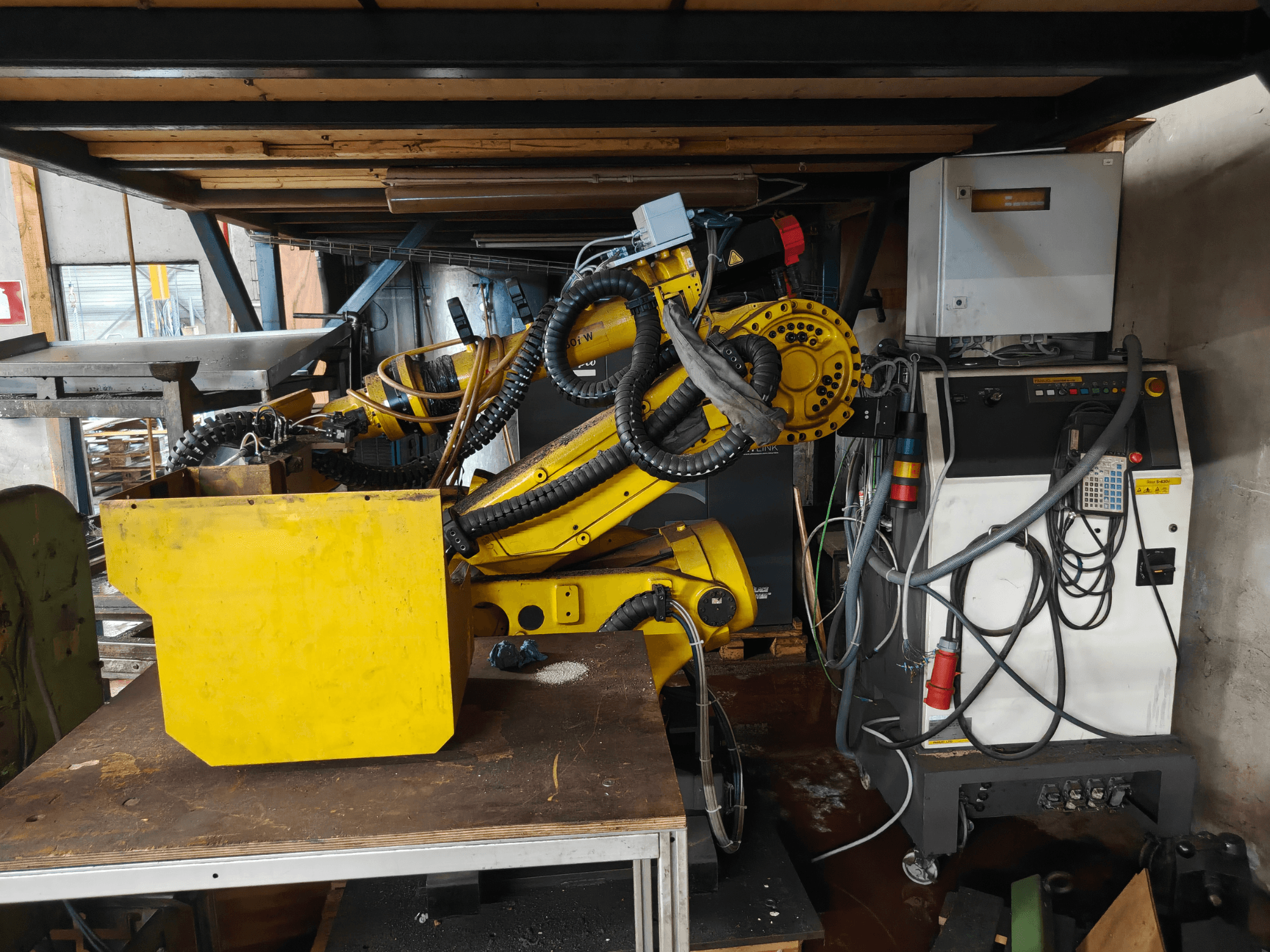 Pohled zepředu na rameno robota FANUC S-430i W, žluté barvy, s ovládacím panelem, kabely a žlutou součástí na pracovním stole.