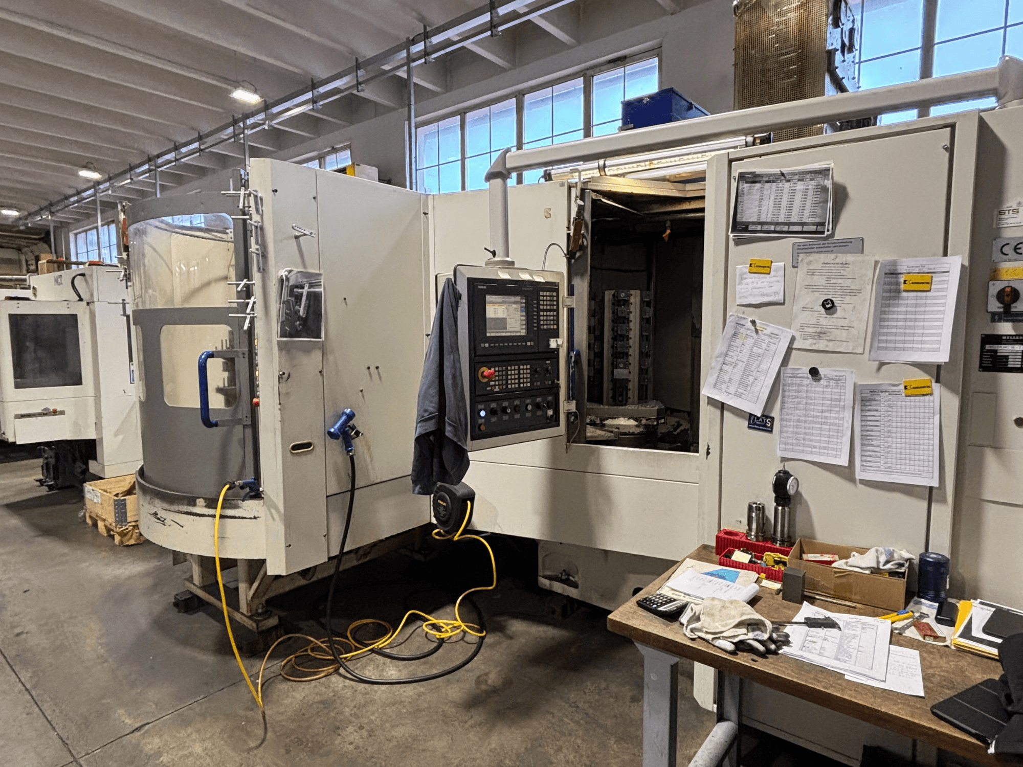 CNC stroj v dílně, pohled zepředu, s ovládacím panelem a dokumentací na boku; nástroje a materiály na stole.