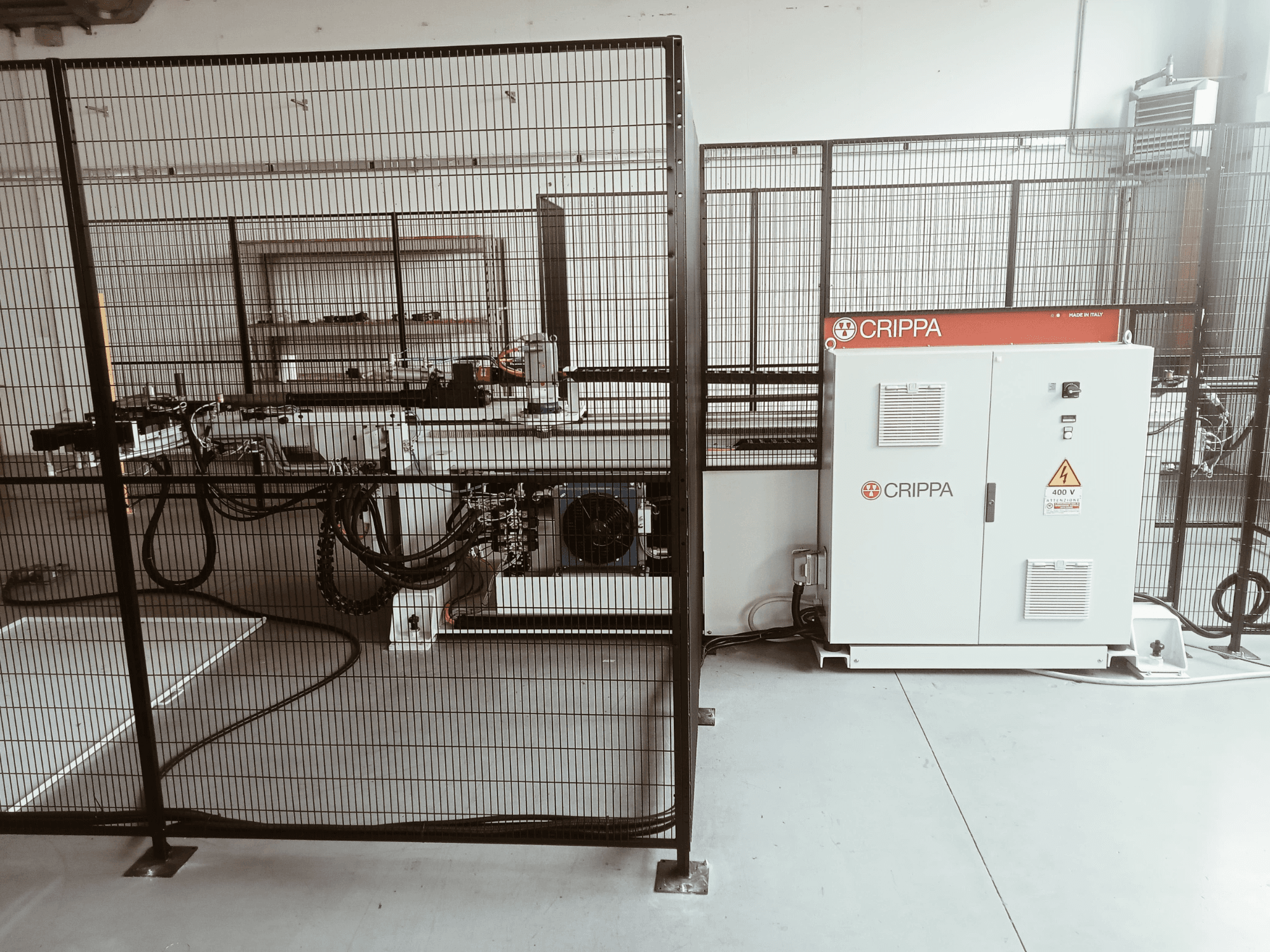 Bílý stroj CRIPPA za bezpečnostní klecí, s viditelnými hadicemi a součástmi, boční pohled, průmyslové prostředí.