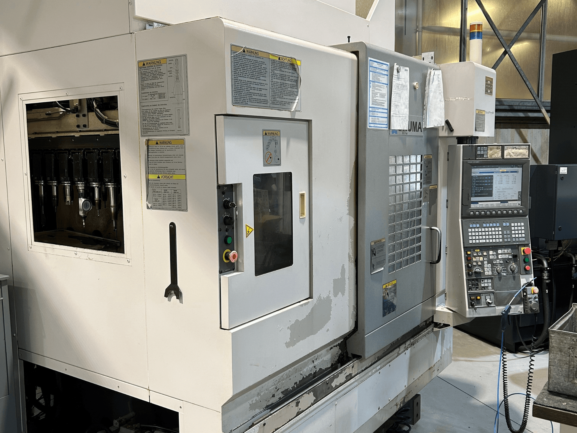 Pohled zepředu na stroj UMA CNC s viditelnými držáky nástrojů, ovládacím panelem, výstražnými štítky a ovládacími tlačítky.