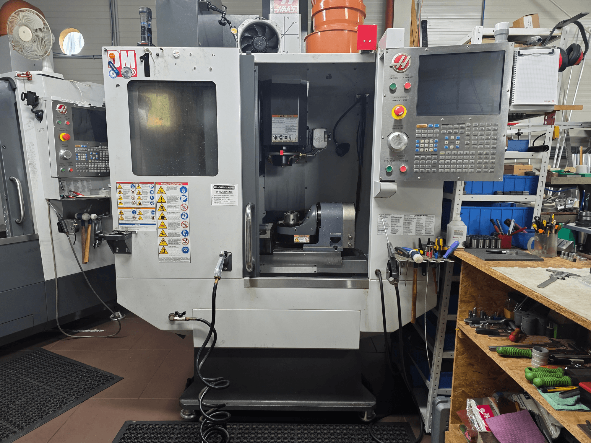 CNC stroj Haas model VF-2, pohled zepředu, zobrazující ovládací prvky, prostor vřetena, bezpečnostní štítky a nástroje na nedalekém pracovním stole.