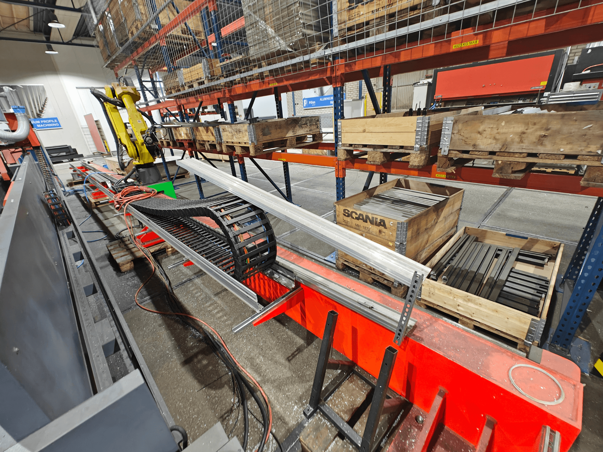 Robotické rameno na pracovišti, viditelné oranžové a černé kabely, s bednami Scania a hliníkovými profily v továrním prostředí.