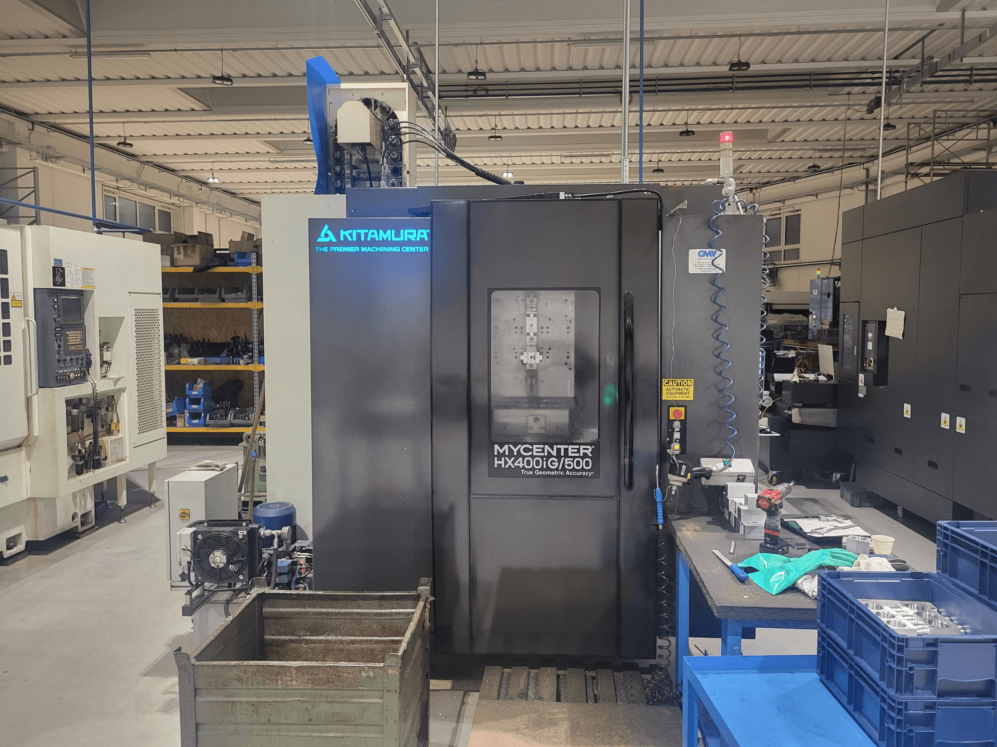 CNC stroj Kitamura Mycenter HX400iG/500 v dílně, pohled zepředu, s ovládacími prvky, úložným prostorem pro nástroje a materiály v blízkosti.
