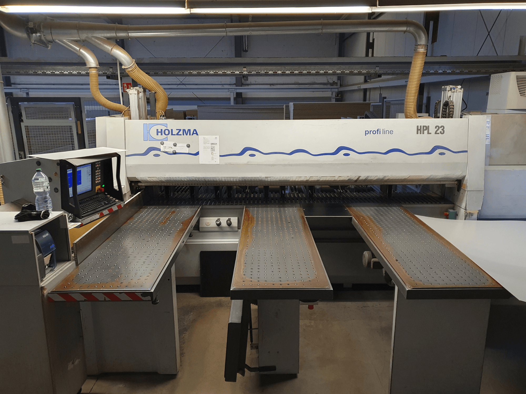 Řezací stroj HOLZMA HPL 23 zobrazený z čelního pohledu, se třemi děrovanými stoly, počítačovým terminálem a lahví s vodou poblíž.