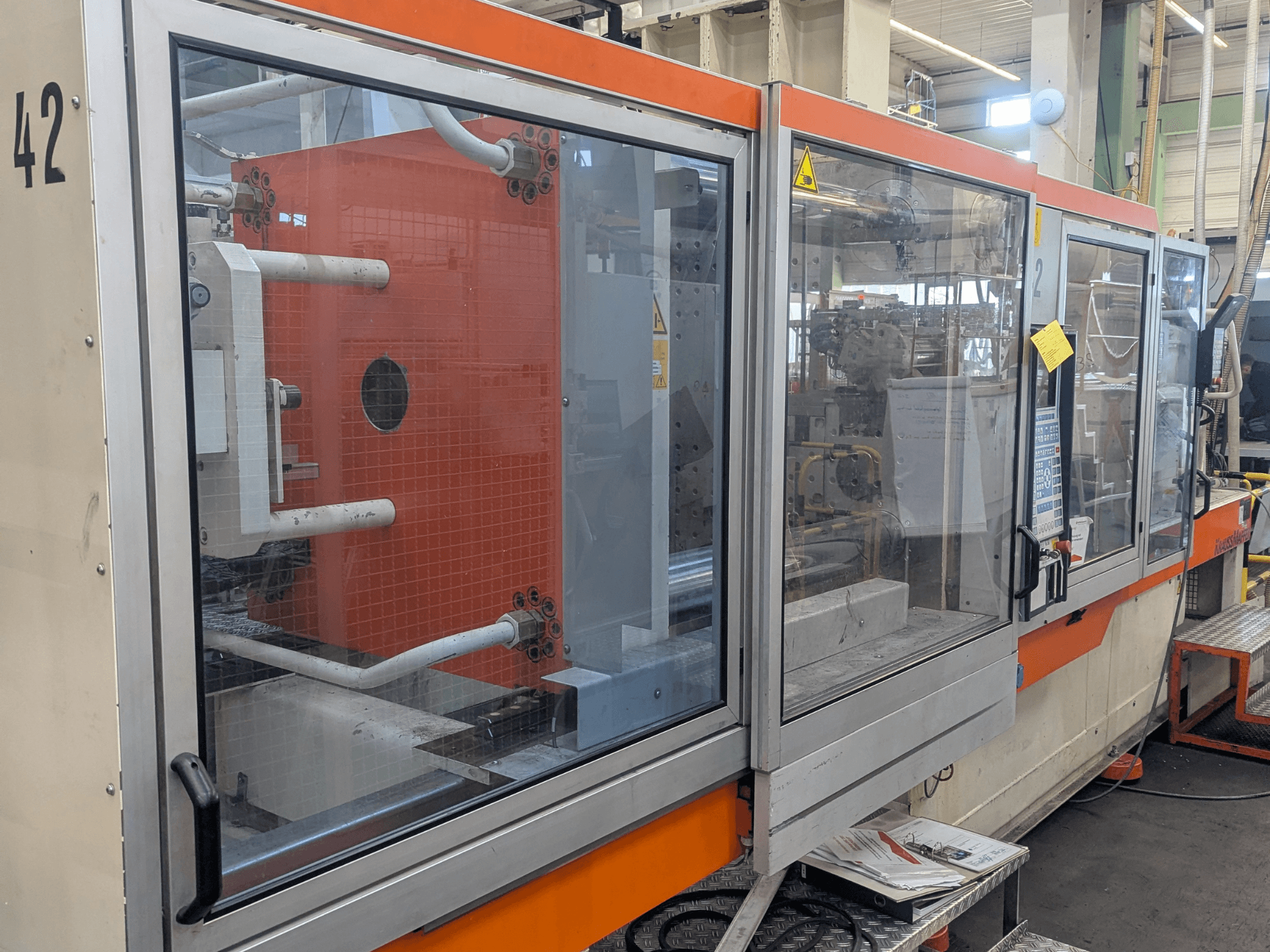 Hydraulický vstřikovací stroj Krauss Maffei KM 350-3000, boční pohled zobrazující ovládací prvky a vnitřní komponenty za skleněným panelem.