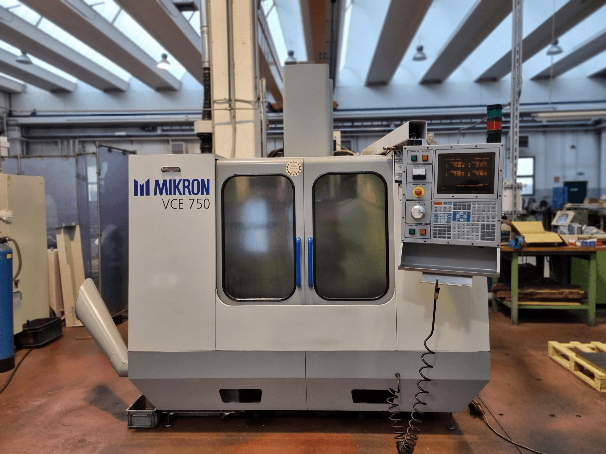 Bílý stroj Mikron VCE 750 CNC v dílně, boční pohled, s digitálním ovládacím panelem a velkými prosklenými dveřmi.