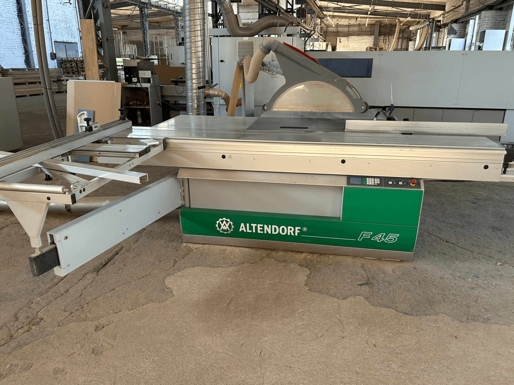 Plošná pila Altendorf F 45, pohled zepředu, na kterém je vidět řezací stůl, kryt pilového kotouče a ovládací panel na stroji v zelené a šedé barvě.