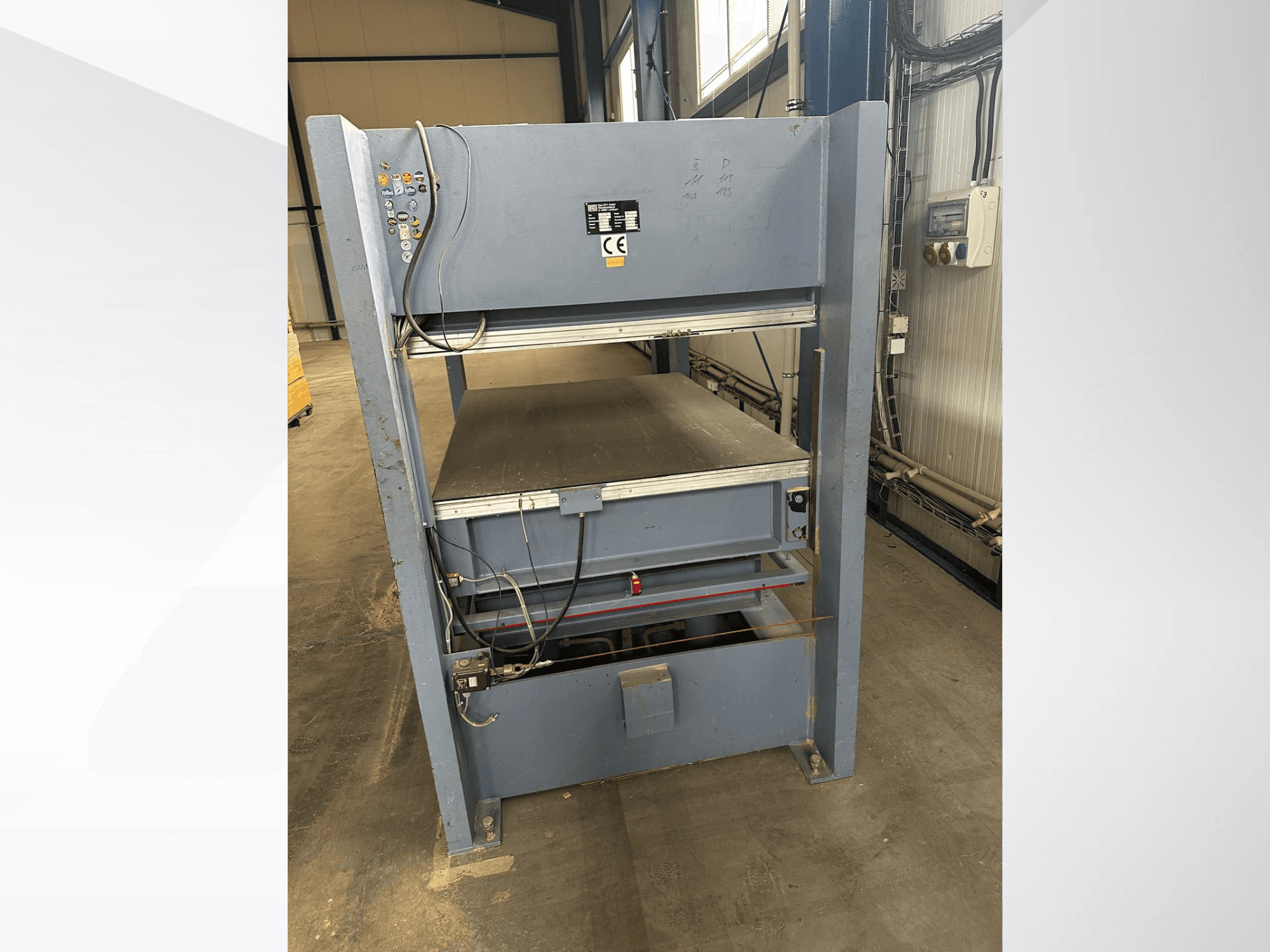 Šedý průmyslový stroj s ovládacím panelem, pohled zepředu; obsahuje nastavitelné plošiny a bezpečnostní značení.