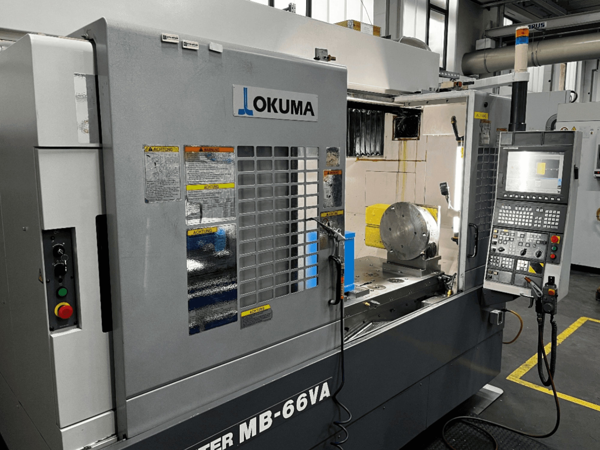 Vertikální obráběcí centrum Okuma MB66-VA z čelního pohledu zobrazující ovládací panel, obrobek a vnitřní prostor stroje s bezpečnostními štítky.