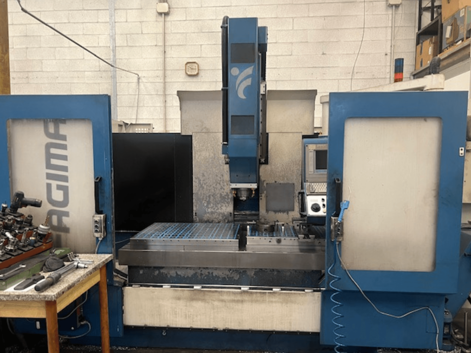 Pohled zepředu na CNC stroj JAGIMA s velkým kovovým pracovním prostorem, viditelným ovládacím panelem a nástroji na vedlejším stole.