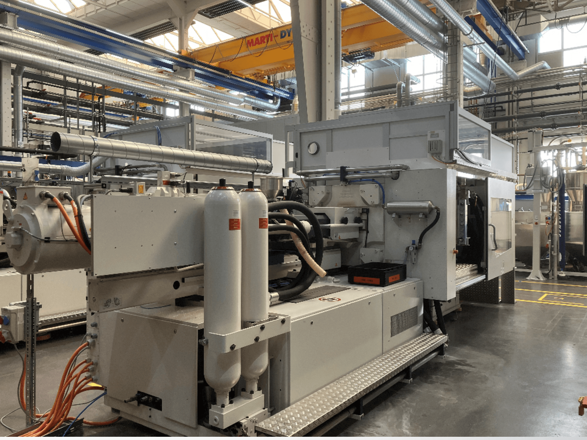 Průmyslový stroj zobrazený z bočního pohledu s několika hadicemi, dvěma vertikálními nádržemi a ovládacím panelem; značka/model není vidět.