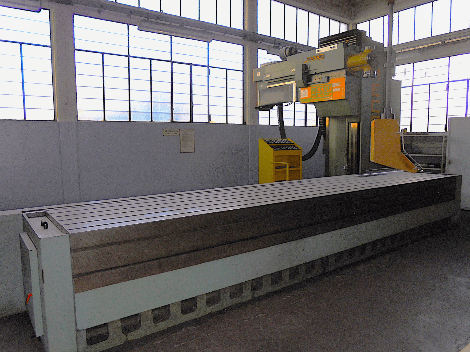 Zcela neznámá vertikální frézka v továrně, pohled z boku, s velkým stolem s drážkovaným povrchem a ovládacím panelem.