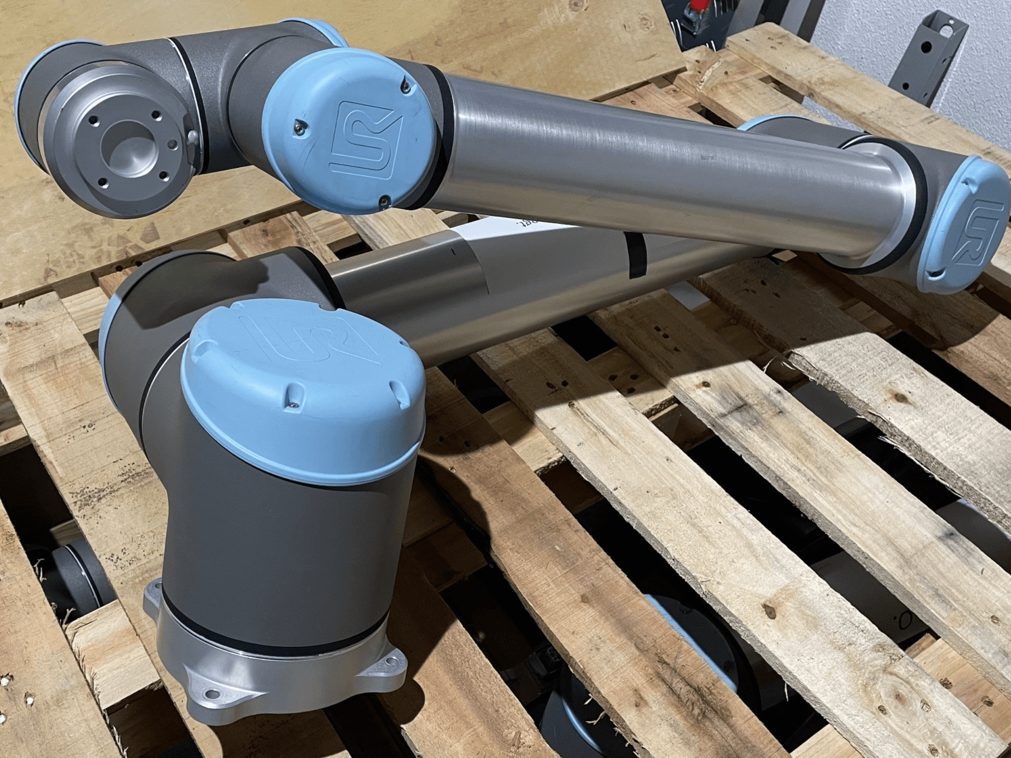 Stříbrná robotická ruka s modrými koncovkami, pohled zepředu, klouby a mechanické detaily, spočívá na dřevěné paletě.