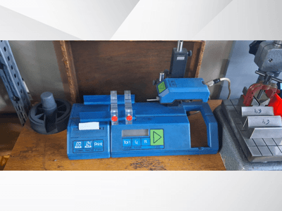 Modrá tiskárna Hommel T 1000 a tester T 1 010 s červenými tlačítky, pohled zepředu na pracovním stole s kovovými bloky.