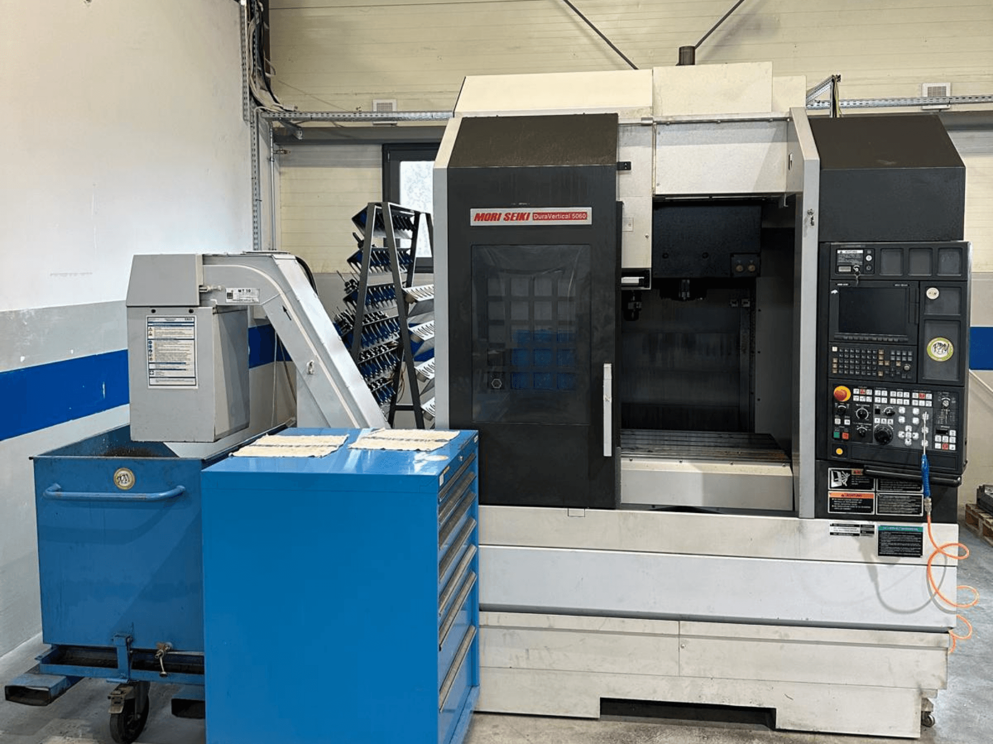 MORI SEIKI DURA VERTICAL 5060, čelní pohled na vertikální obráběcí centrum s ovládacím panelem a přilehlým úložištěm nástrojů.