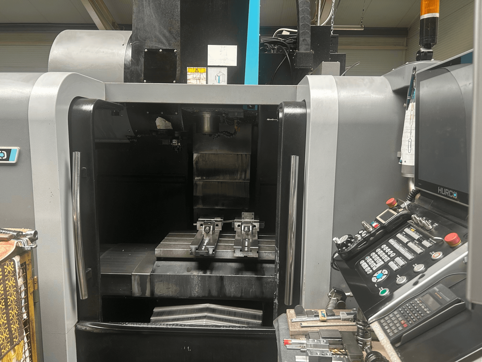 Pohled zepředu na CNC obráběcí centrum Hurco, zobrazující držák nástrojů, kovový obrobek a ovládací panel s tlačítky a displejem.