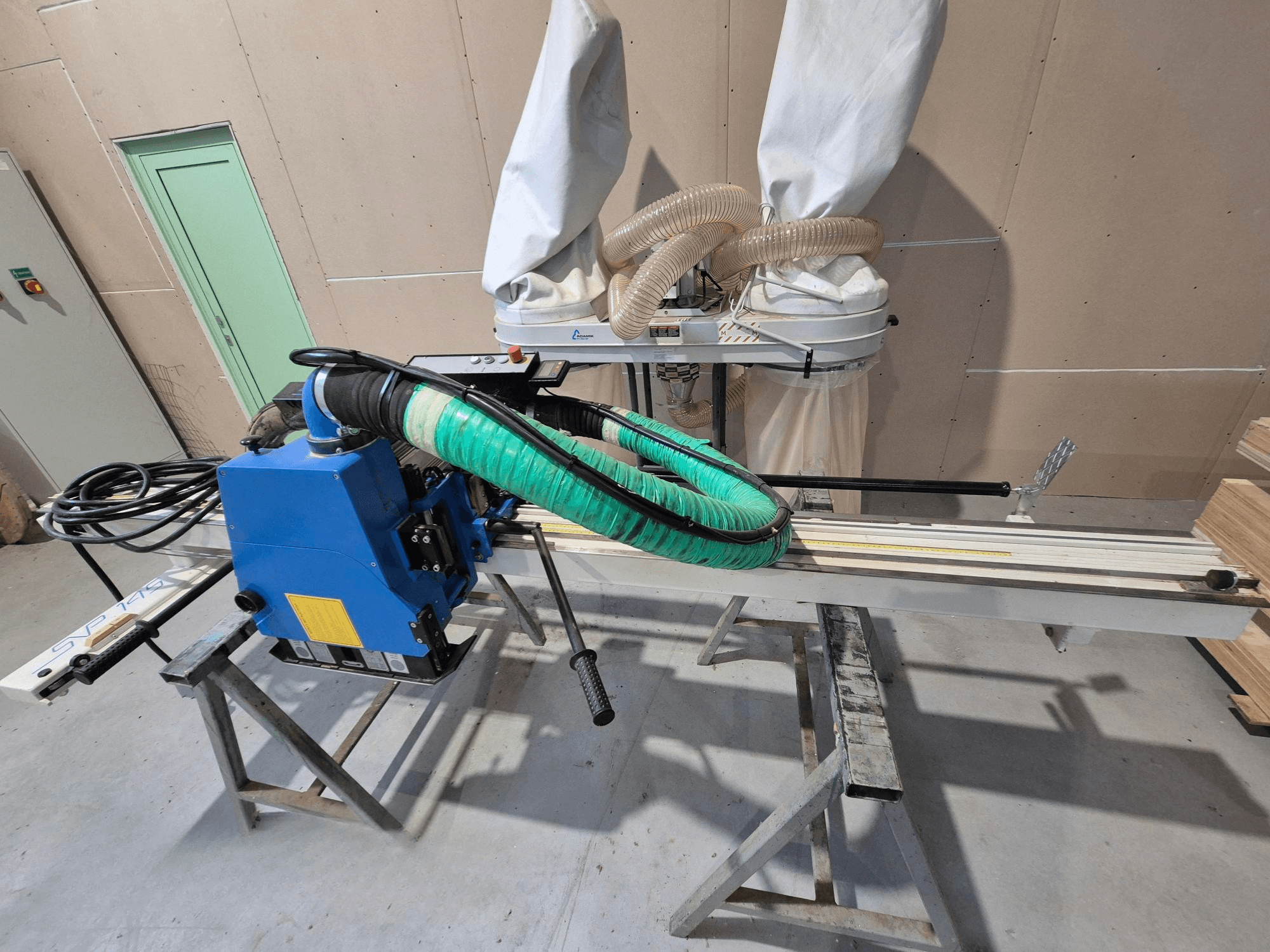Modrá horizontální pila SVP-175 se zelenou prachovou hadicí a podpěrou, pohled z boku v dílně.