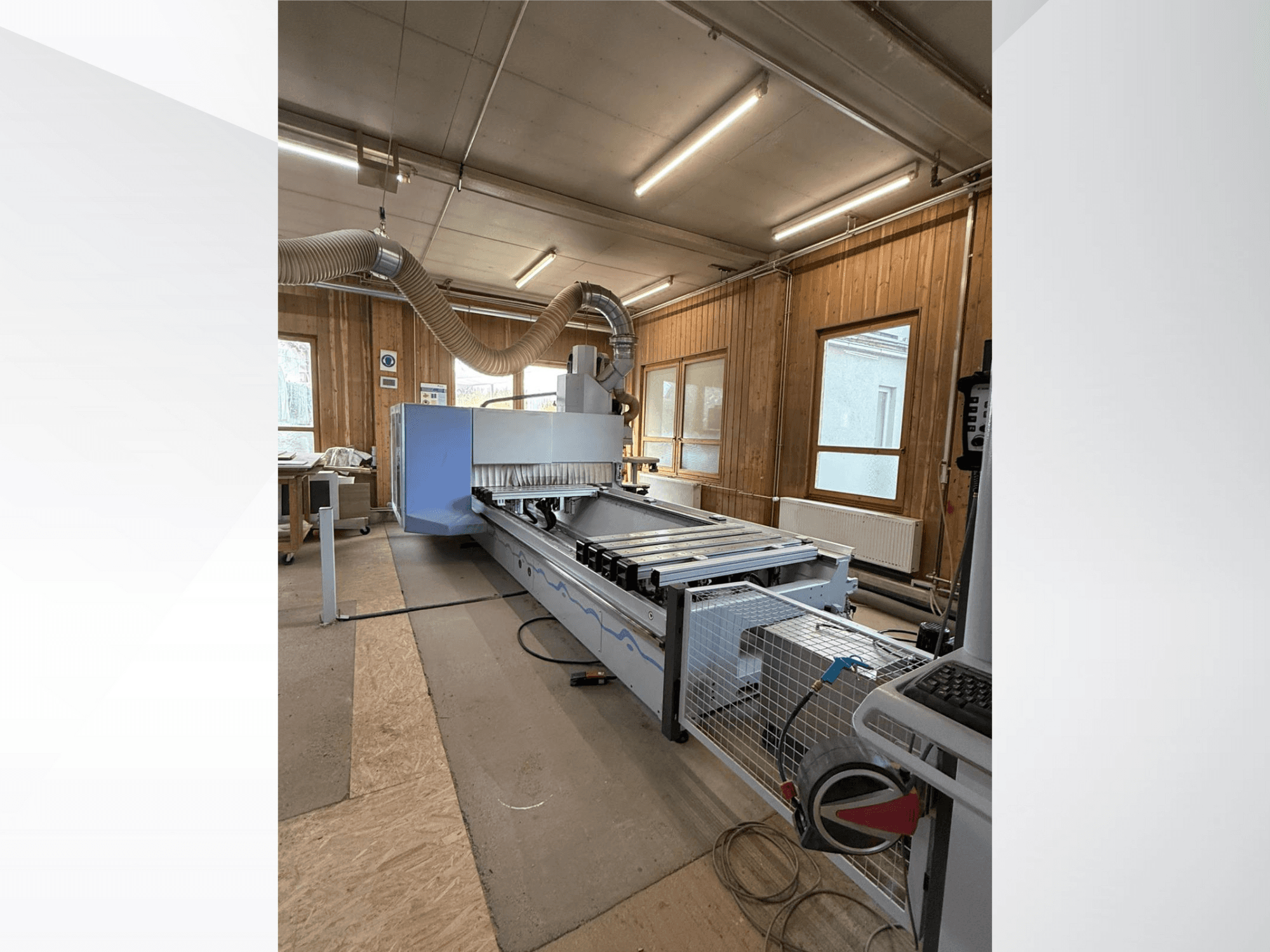 CNC frézka v dílně, boční pohled, s velkým modrým tělesem, připojeným pásovým dopravníkem a viditelným ovládacím panelem.