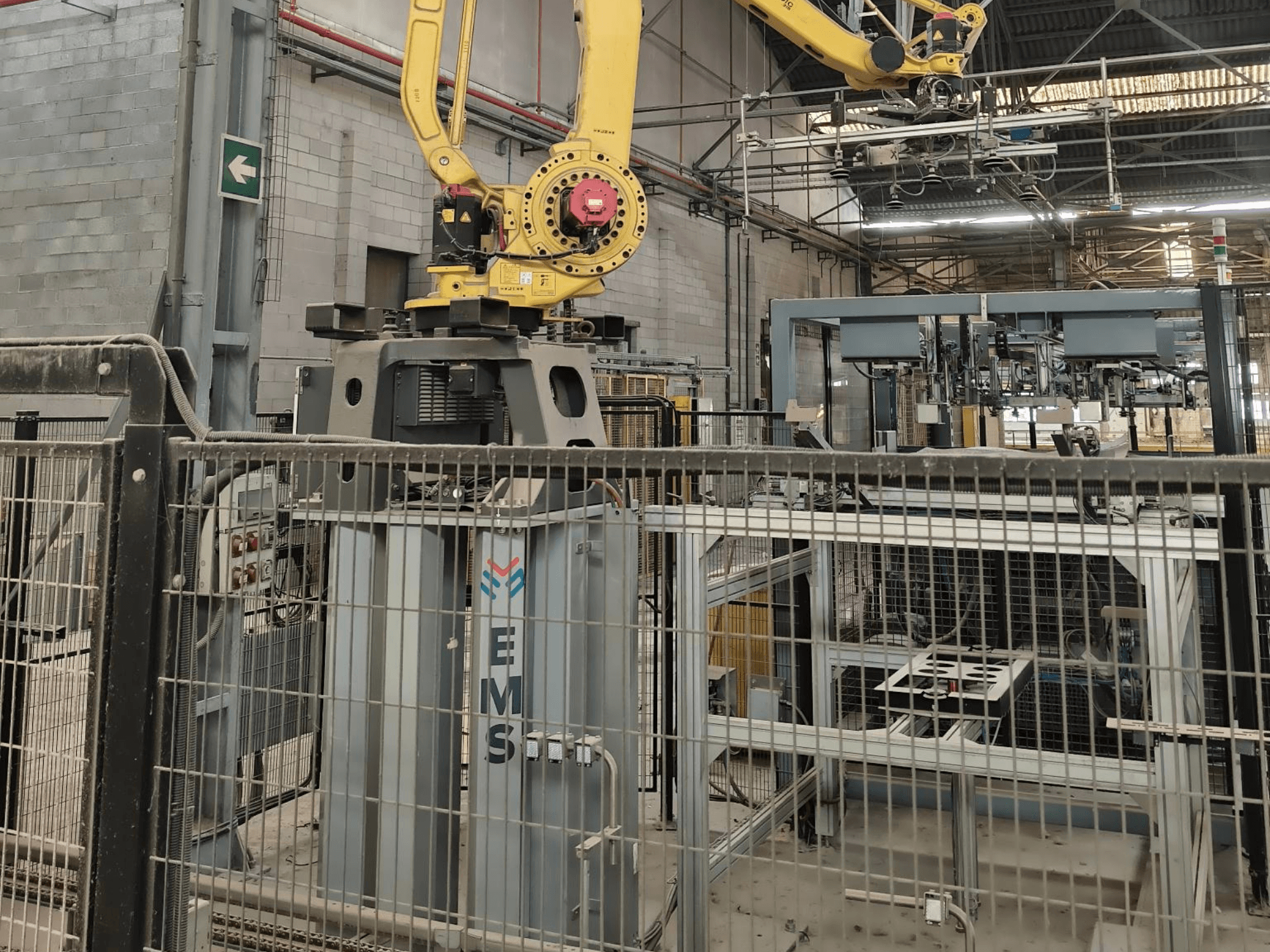 Pohled zepředu na robotické rameno FANUC M-410iC/315 umístěné nad nástrojem EMS, v pozadí je vidět průmyslové zařízení a ochranné oplocení.