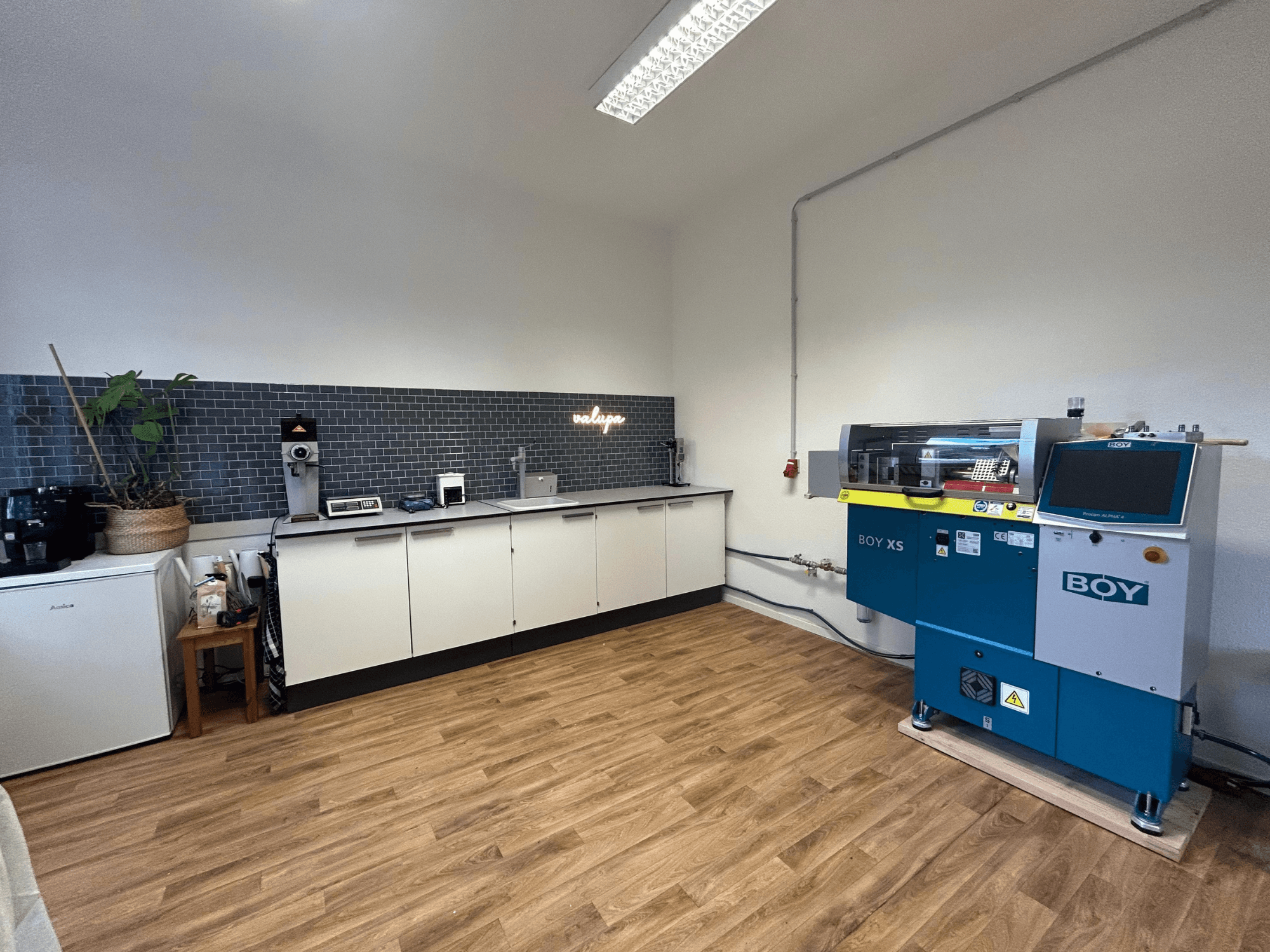 Hydraulický vstřikovací stroj BOY XS 161, pohled zepředu, v moderní kuchyni se spotřebiči a dřevěnou podlahou.