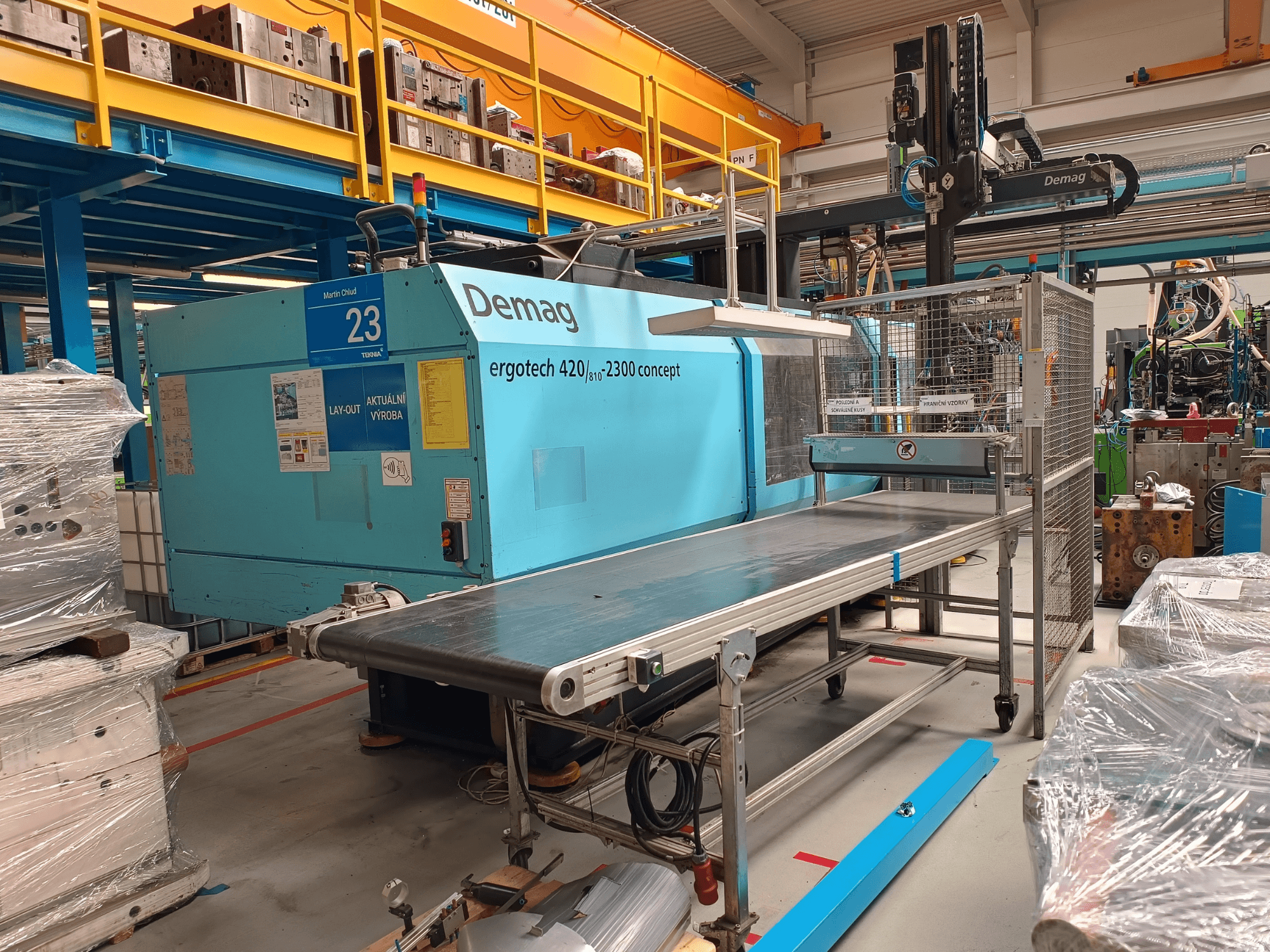 Hydraulický vstřikovací stroj Demag ergotech 420/810 - 2300 v bočním pohledu, s dopravníkem a bezpečnostním krytem.