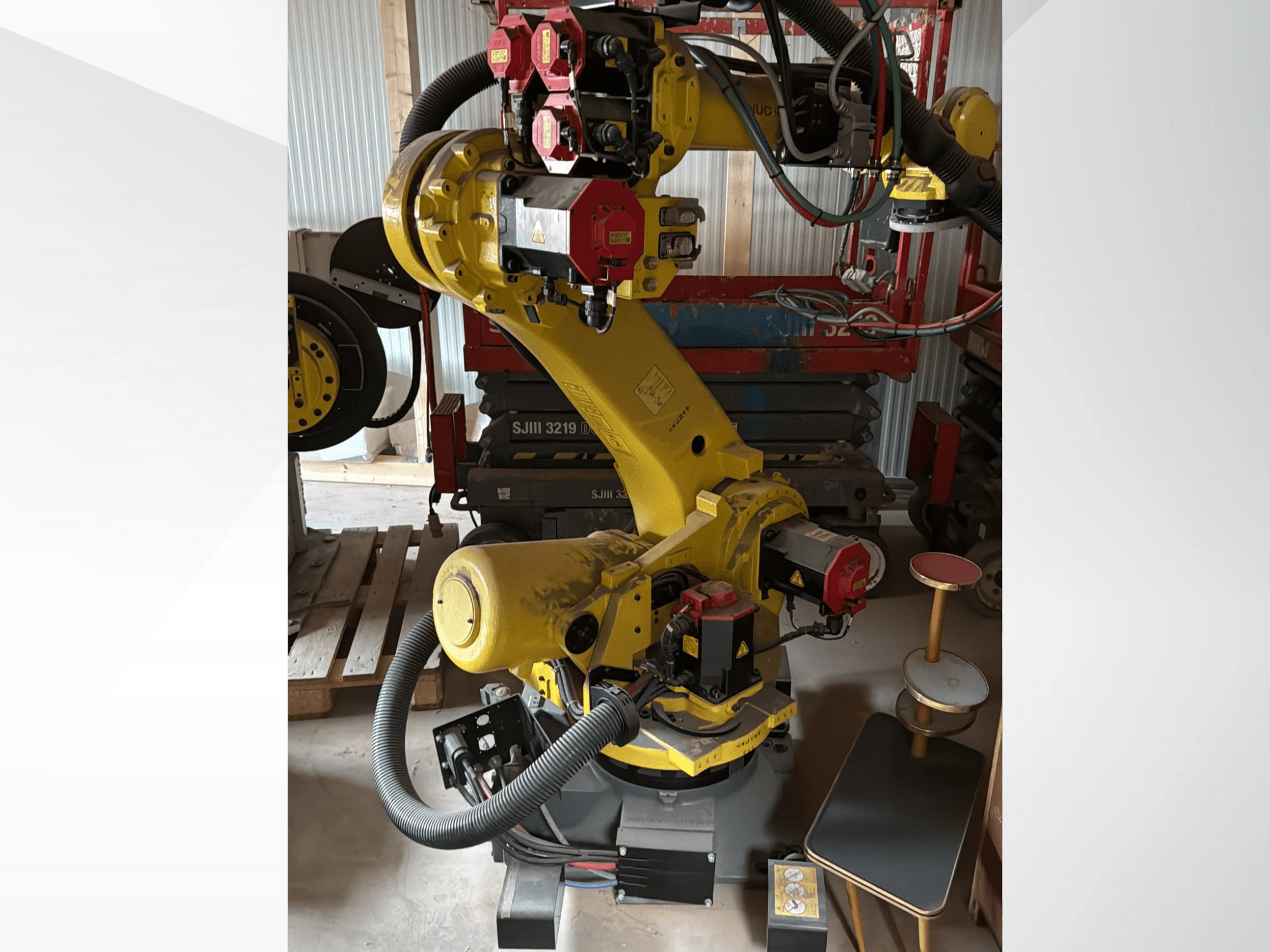 Žluté průmyslové robotické rameno s červenými ovladači, pohled zepředu; s několika klouby a hadicemi, umístěné na pracovišti.