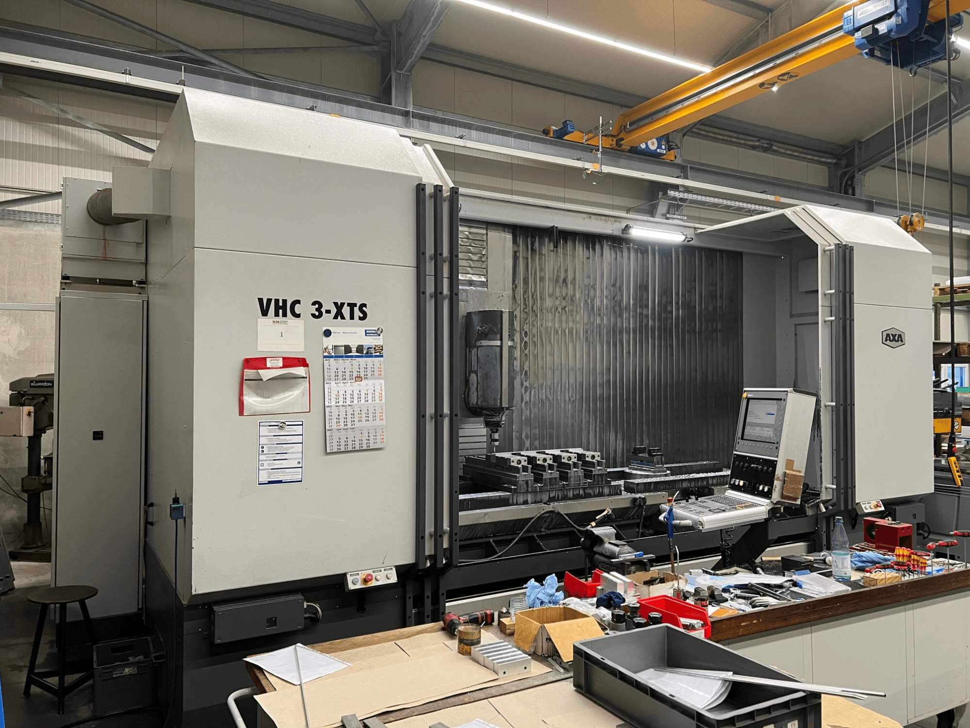 CNC stroj AXA VHC 3-XTS v bočním pohledu, s ovládacím panelem a pracovním prostorem přeplněným nástroji a součástmi.