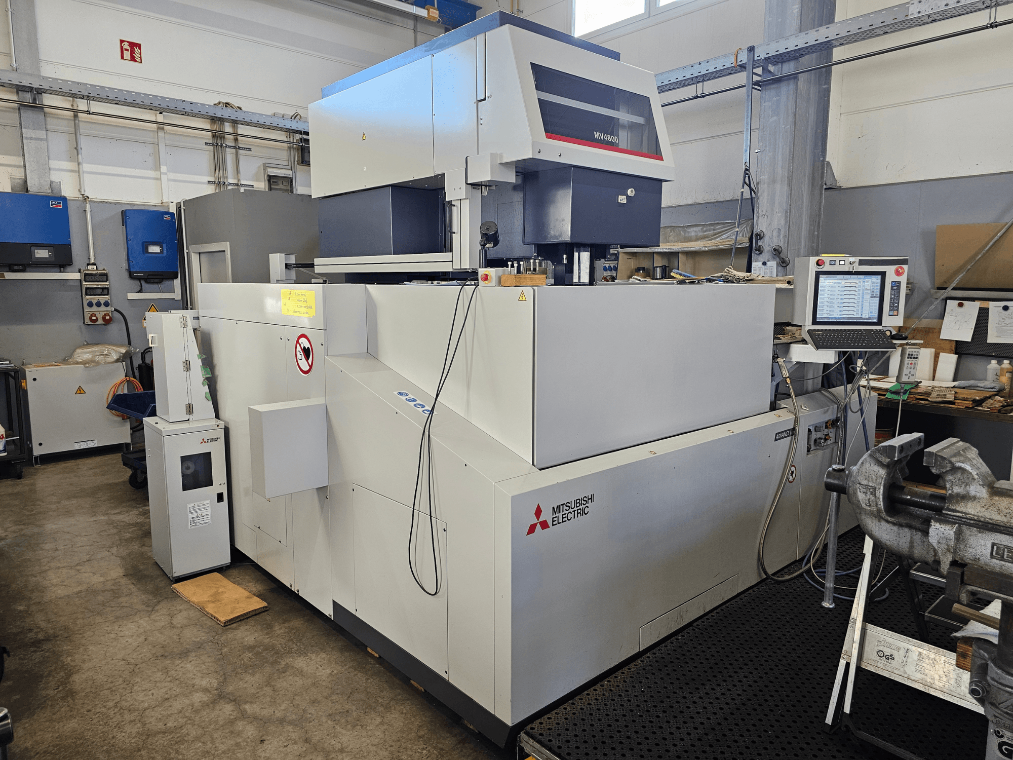 Drátový elektroerozivní stroj Mitsubishi MV4800 v dílně, boční pohled ukazuje ovládací panel, bezpečnostní štítky a okolní vybavení.