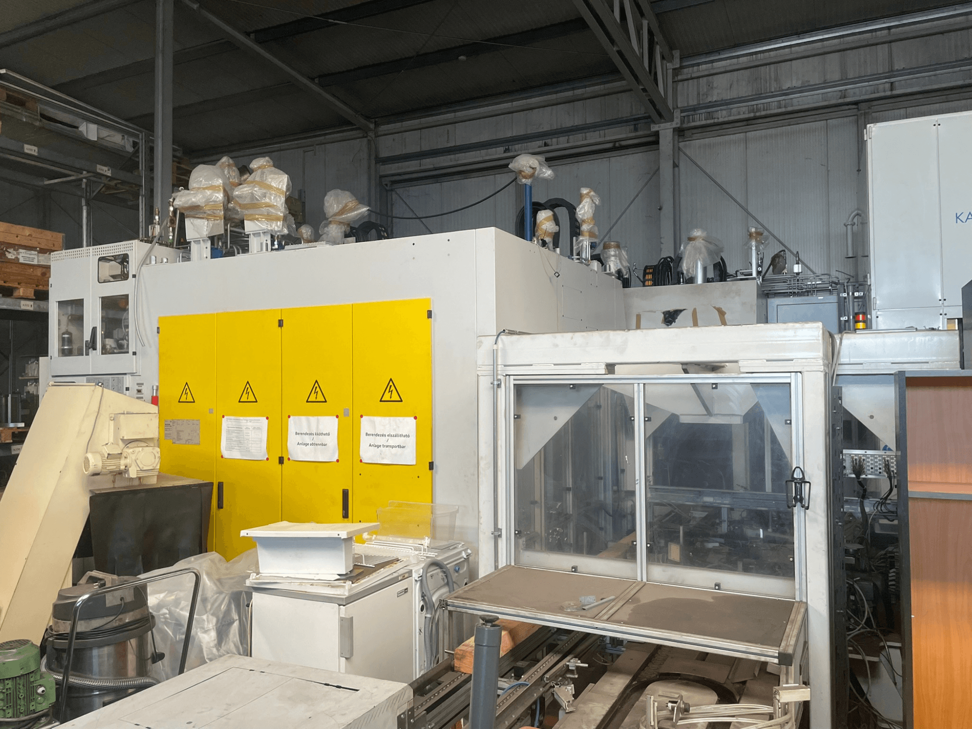 Průmyslové stroje se žlutým ovládacím panelem, pohled z boku, s výstražnými štítky a plastovými kryty, ve skladu.