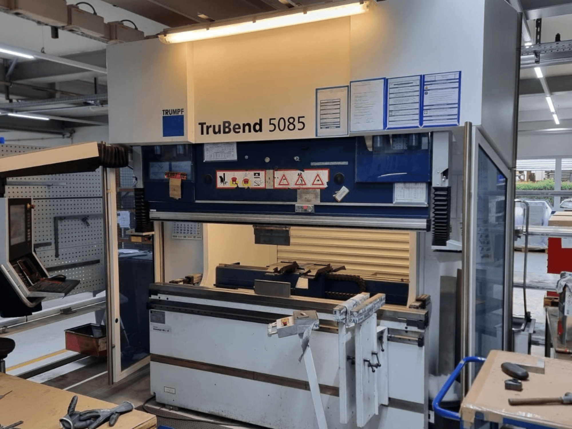 Ohýbačka TRUMPF TruBend 5085, pohled zepředu, zobrazující ovládací panel, obslužné místo a bezpečnostní upozornění uvnitř dílny.