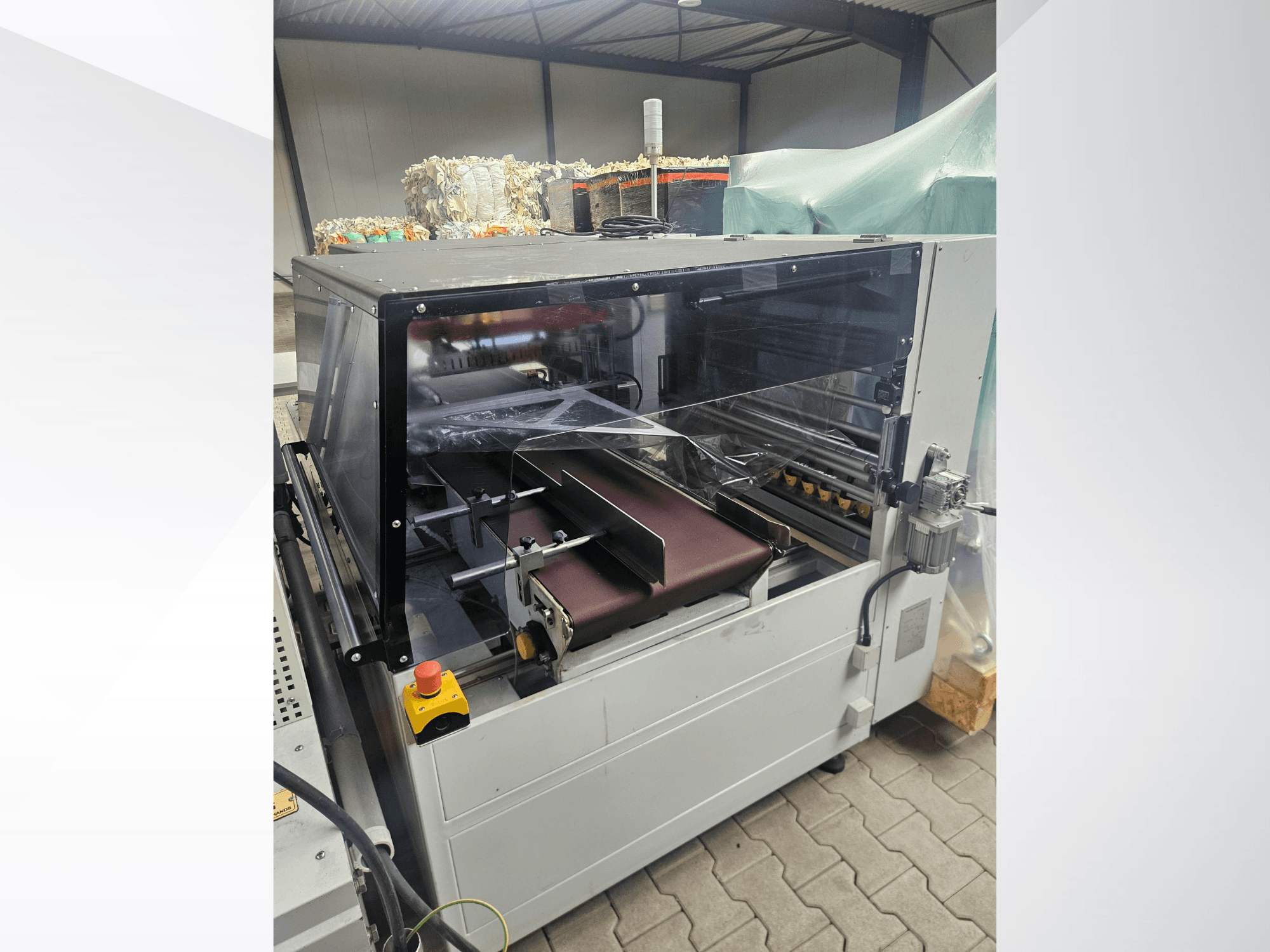Stroj AMTEC Speed Shrink 40 PRO, pohled zepředu, znázorňující mechanismus pro zpracování plastů s průhledným bezpečnostním krytem a dopravním pásem.