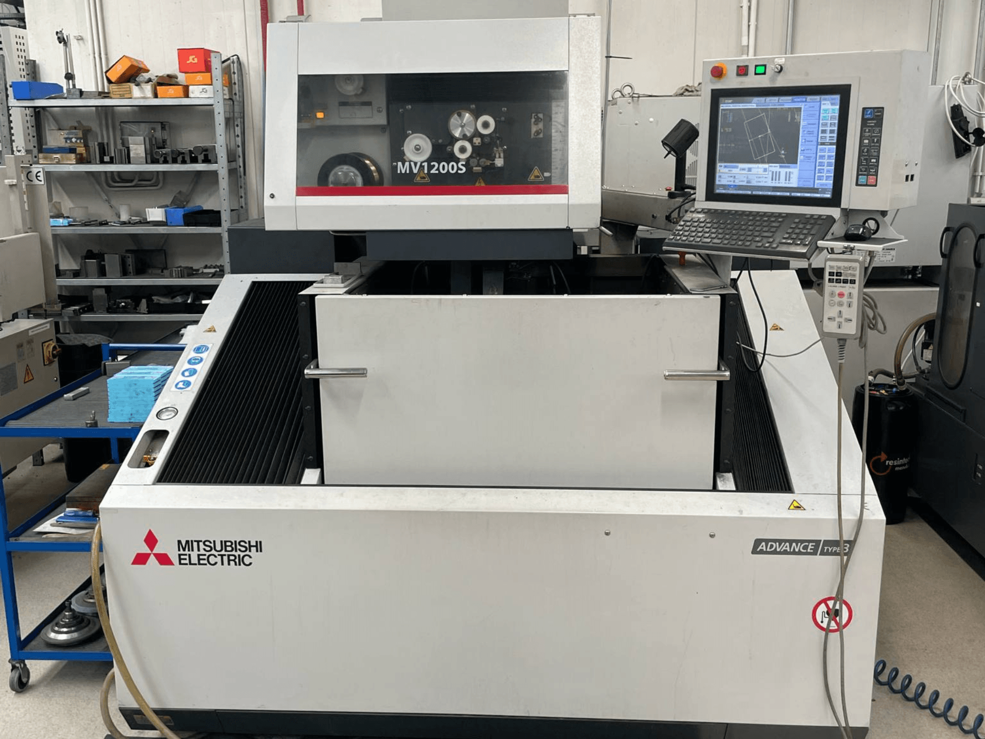 Elektroerozivní stroj Mitsubishi MV1200S v čelním pohledu, na kterém je vidět ovládací panel a pracovní prostor, včetně viditelných bezpečnostních upozornění.