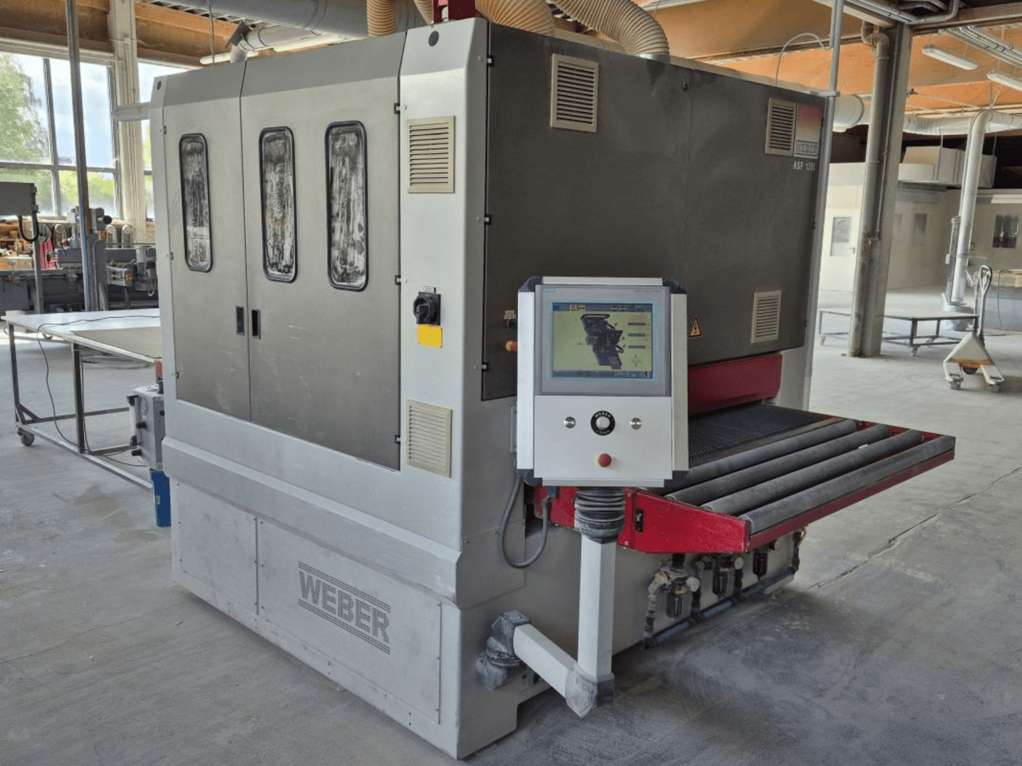 Pásová bruska WEBER KSF-3-1350, boční pohled na ovládací prvky, napájecí panel a brusný dopravník. Průmyslové prostředí s viditelnými stroji.