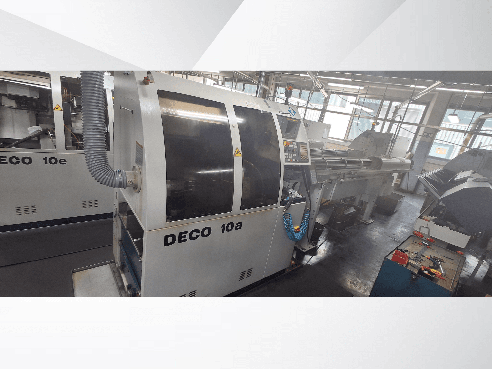 CNC stroj DECO 10a v dílně, pohled zepředu, ovládací panel a výfukový systém. Vyznačuje se robustní konstrukcí a přesnými nástroji.