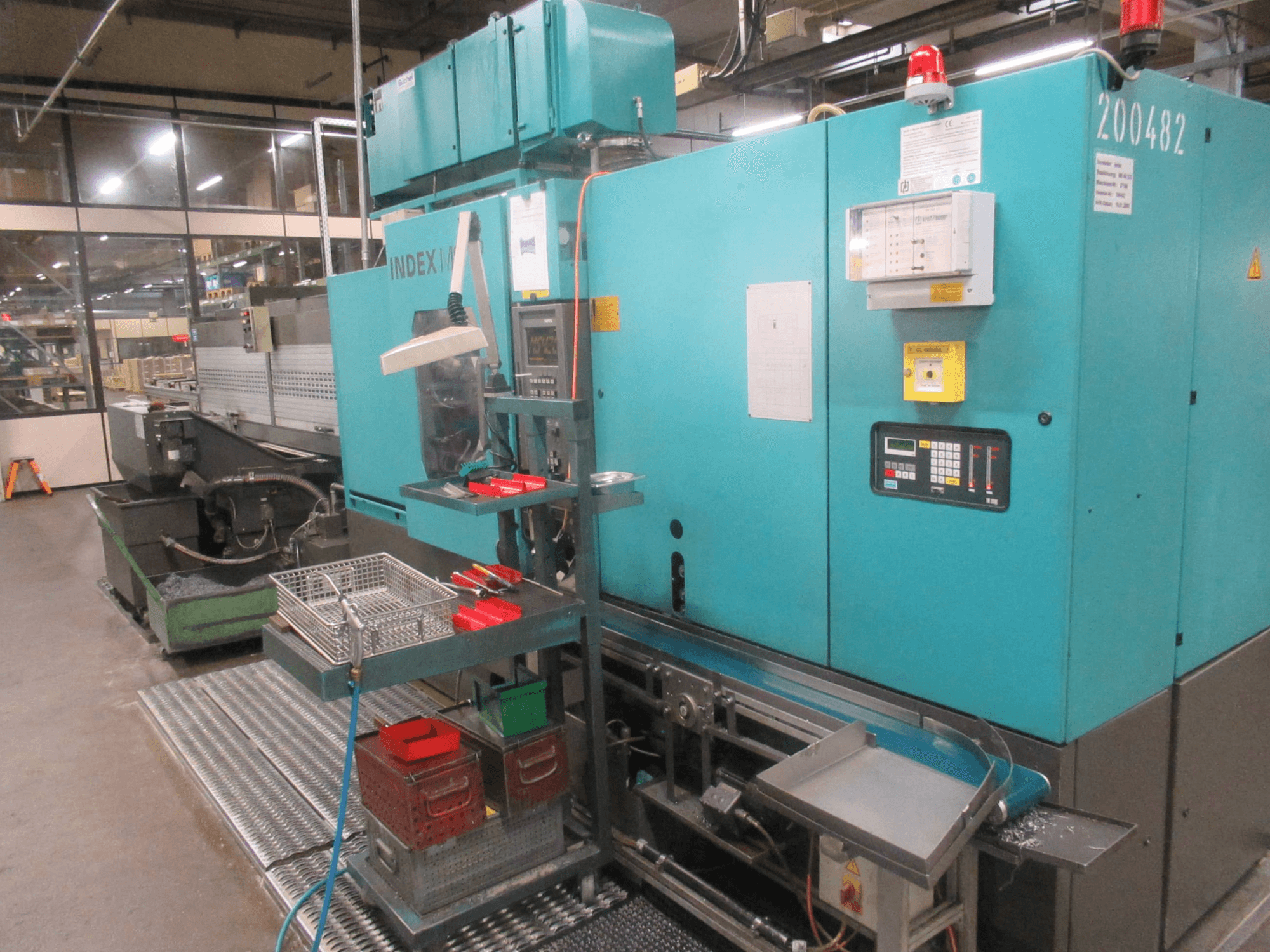 Tyrkysový stroj INDEX v továrně, boční pohled; ovládací panel, zásobník na nástroje a dopravník. Viditelné zásobníky, hadice a alarmy.