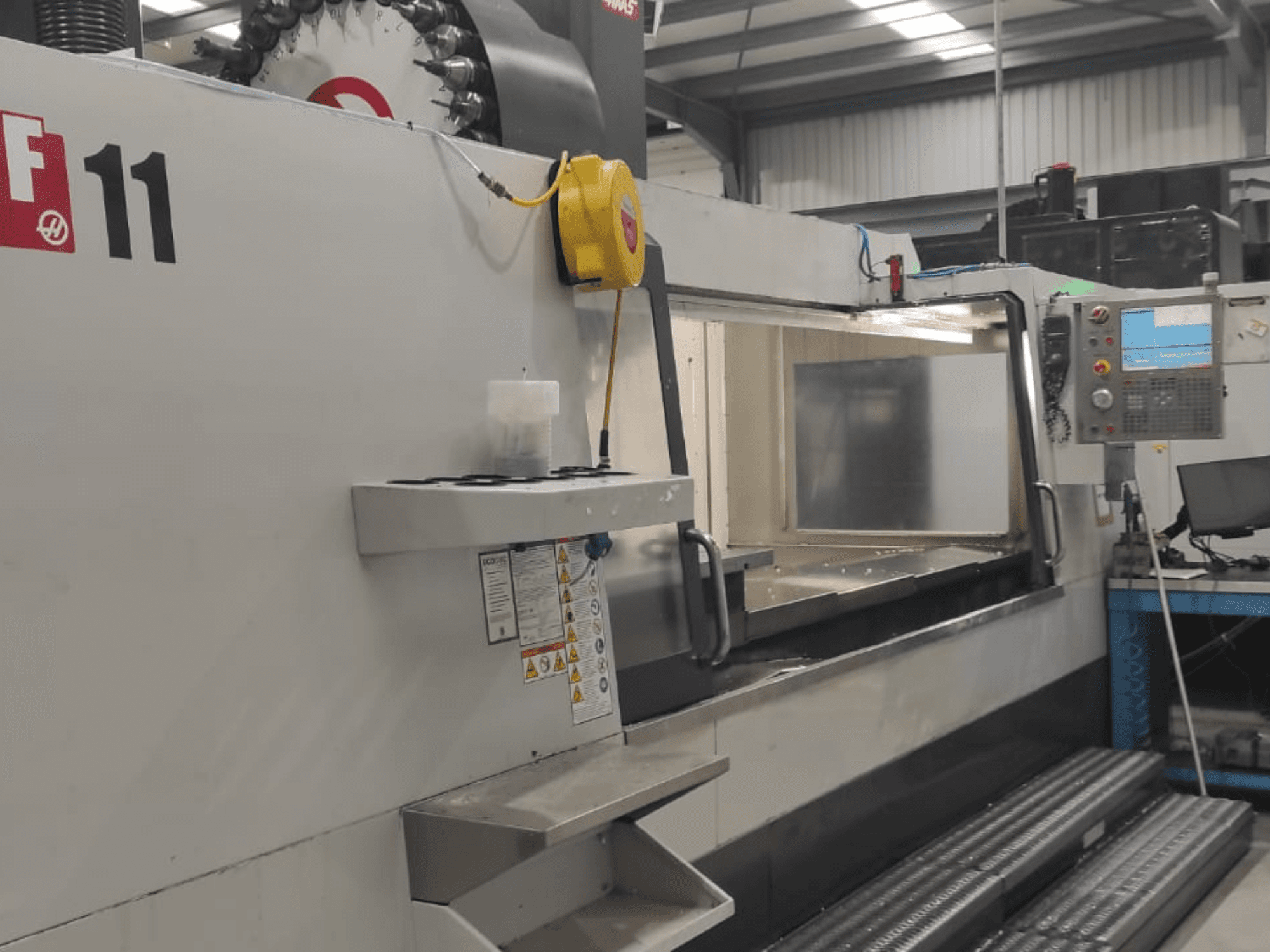 Stroj od společnosti Haas, model VF-11, zobrazený z čelního pohledu s viditelným ovládacím panelem, velkou pracovní plochou a bezpečnostními štítky.