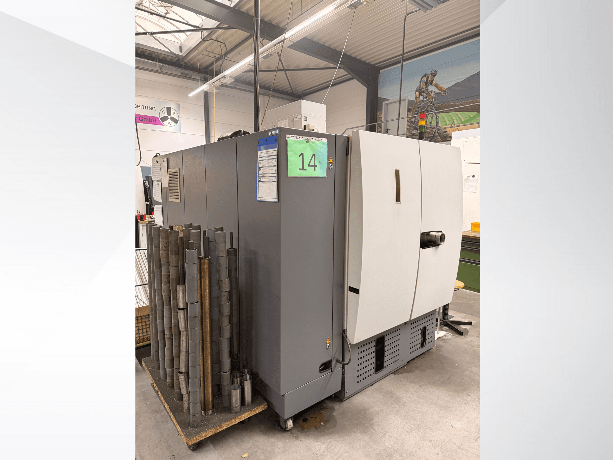 Šedý průmyslový stroj Siemens, boční pohled, vedle něj zásoba válcových kusů kovu a viditelné štítky zařízení.