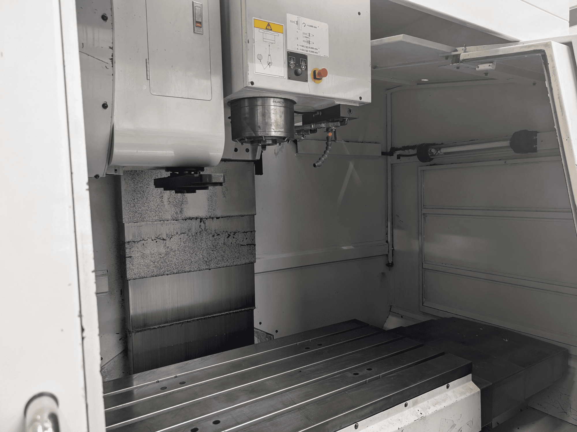 Pohled na CNC frézku s velkým kovovým pracovním stolem a vřetenovým motorem zevnitř, zobrazující detailní komponenty a ovládací panel.