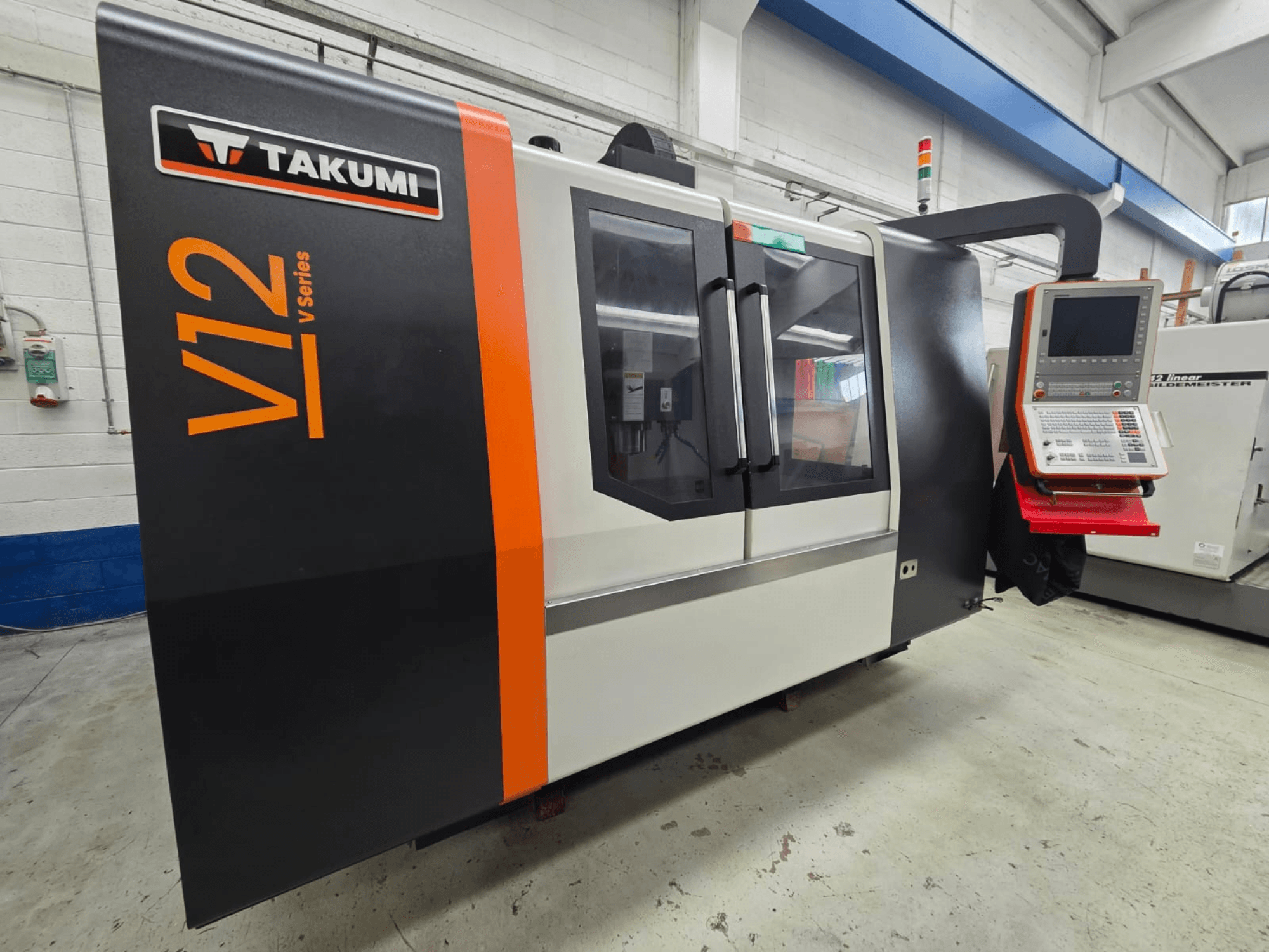 CNC frézka Takumi V12 řady V, pohled zepředu, s ovládacím panelem a velkými skleněnými okny.