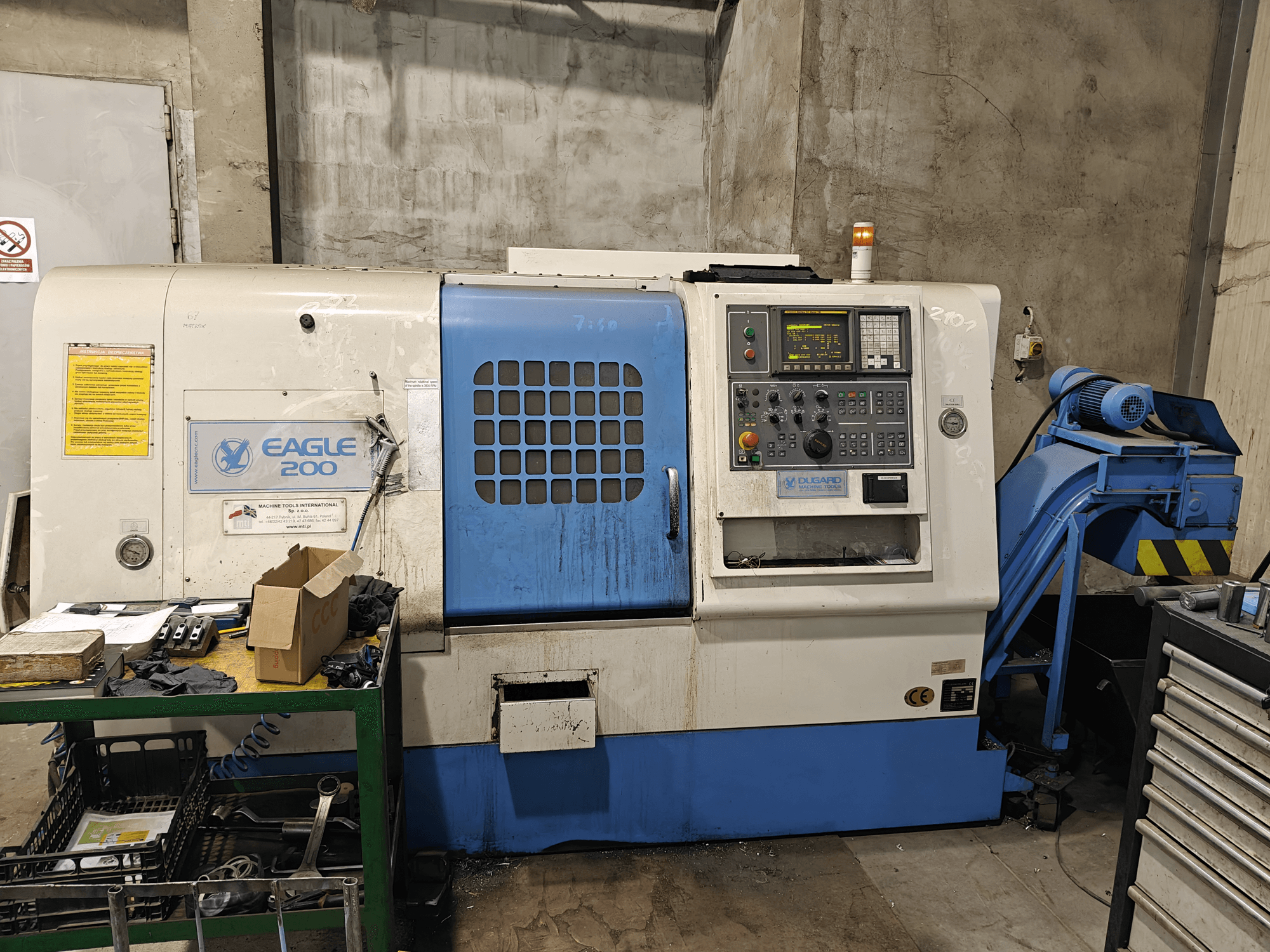 Stroj Eagle 200 CNC, čelní pohled s ovládacím panelem, různé nástroje na zeleném vozíku. Viditelné opotřebení a provozní světla.