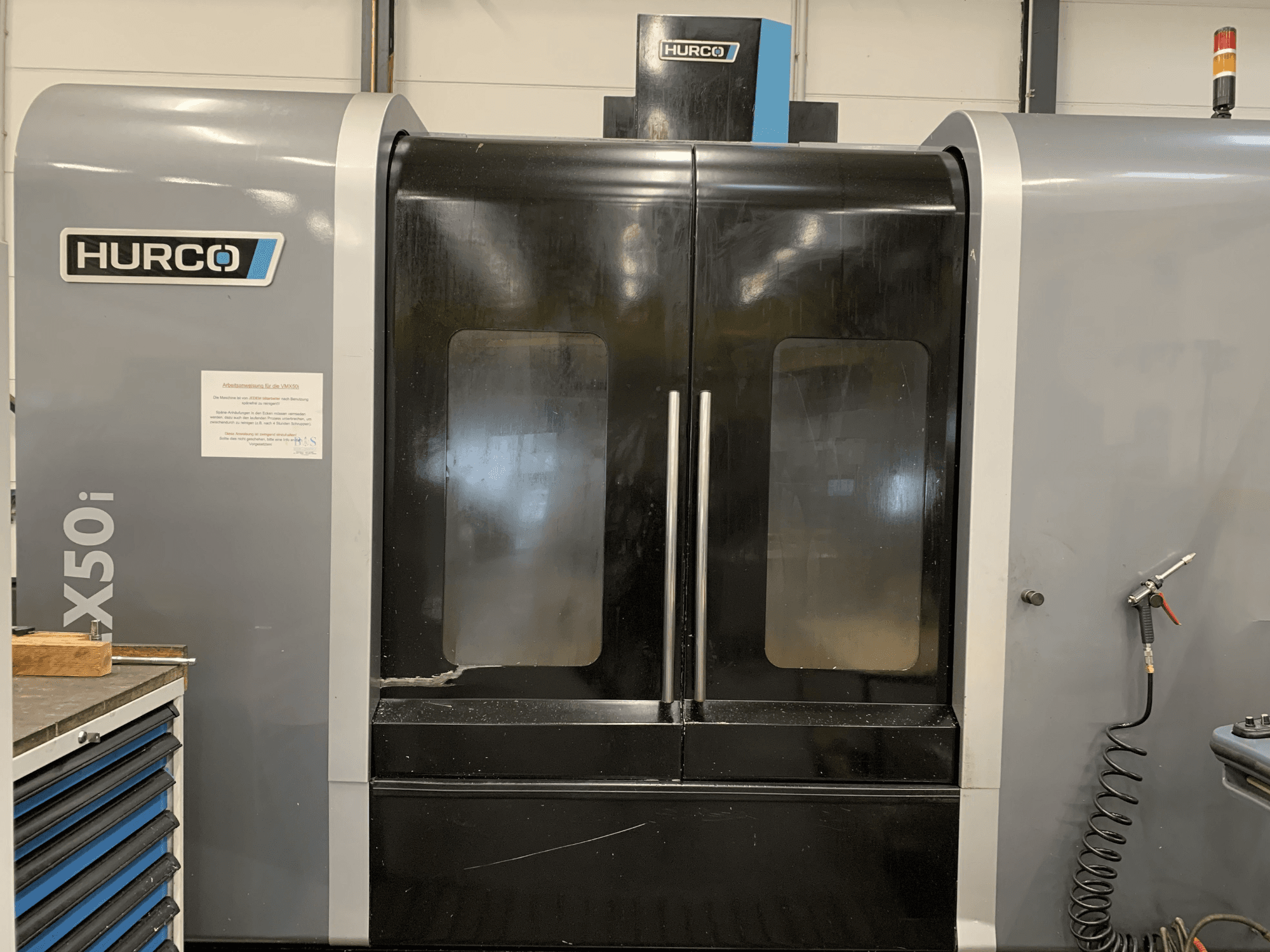 Pohled zepředu na stroj Hurco VX50i CNC se dvěma dveřmi, na kterých jsou vidět technické ovládací prvky a značka údržby na boku.