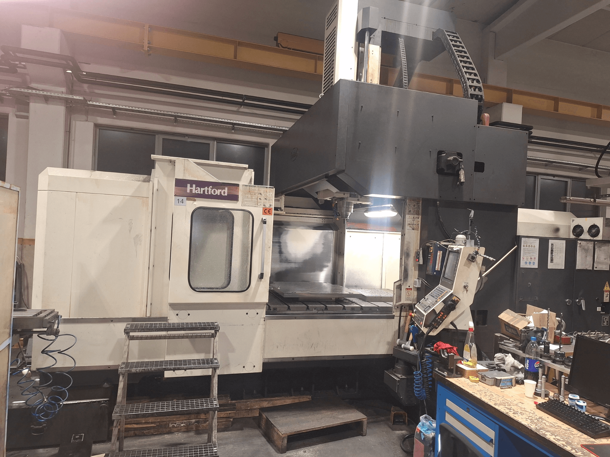 Stroj Hartford CNC, pohled zepředu, s technickými ovládacími prvky, přístupovými schůdky a plochou pracovního stolu v dílně.