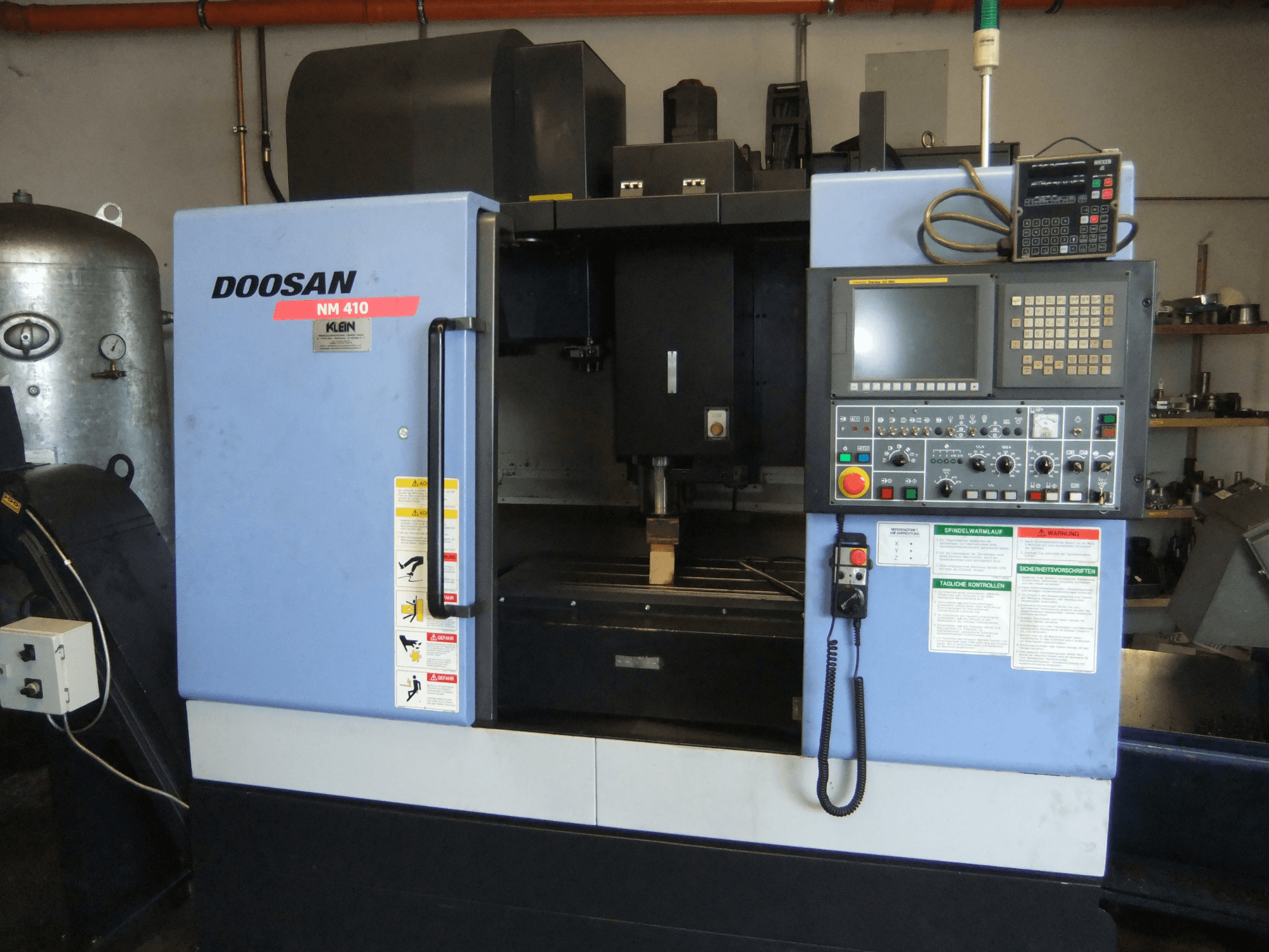 Vertikální obráběcí centrum DOOSAN NM 410, pohled zepředu, na kterém je vidět ovládací panel, oblast vřetena a bezpečnostní štítky.