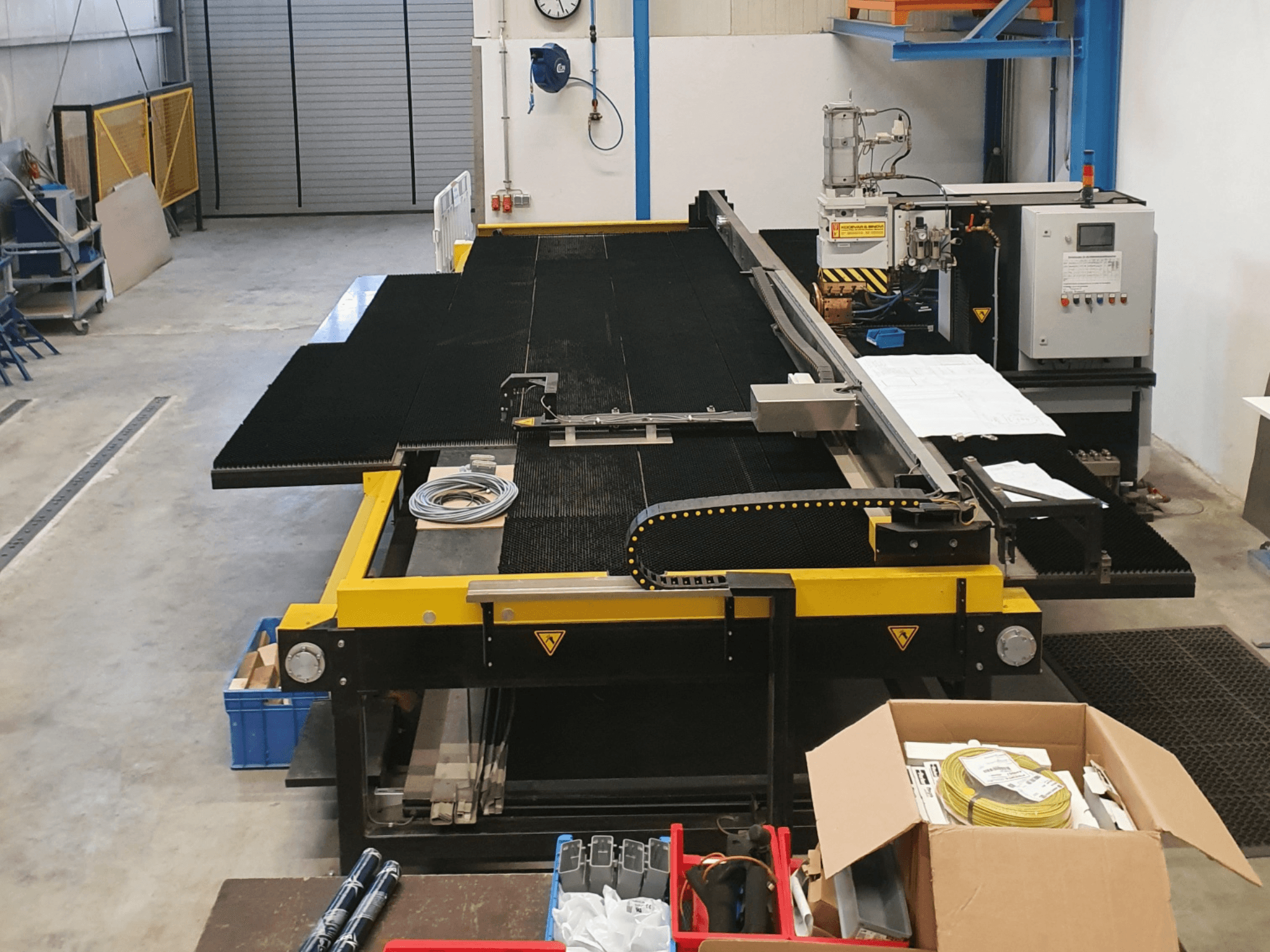 Průmyslový řezací stroj se žlutými a černými prvky, pohled zepředu, zobrazující ovládací prvky a příslušenství v továrním prostředí.