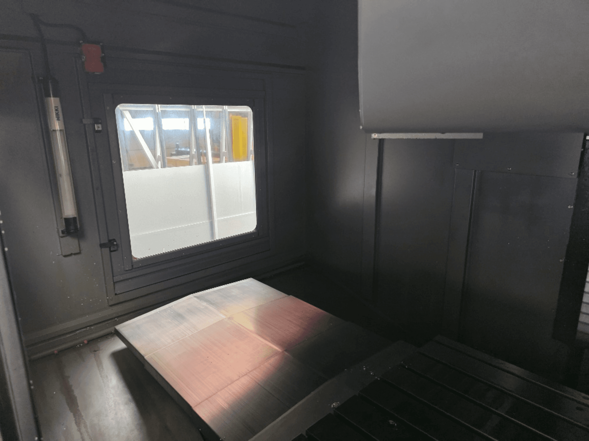 Pohled do interiéru skříně stroje s leštěnou kovovou deskou, bočním oknem a minimálním osvětlením. Značka není viditelná.