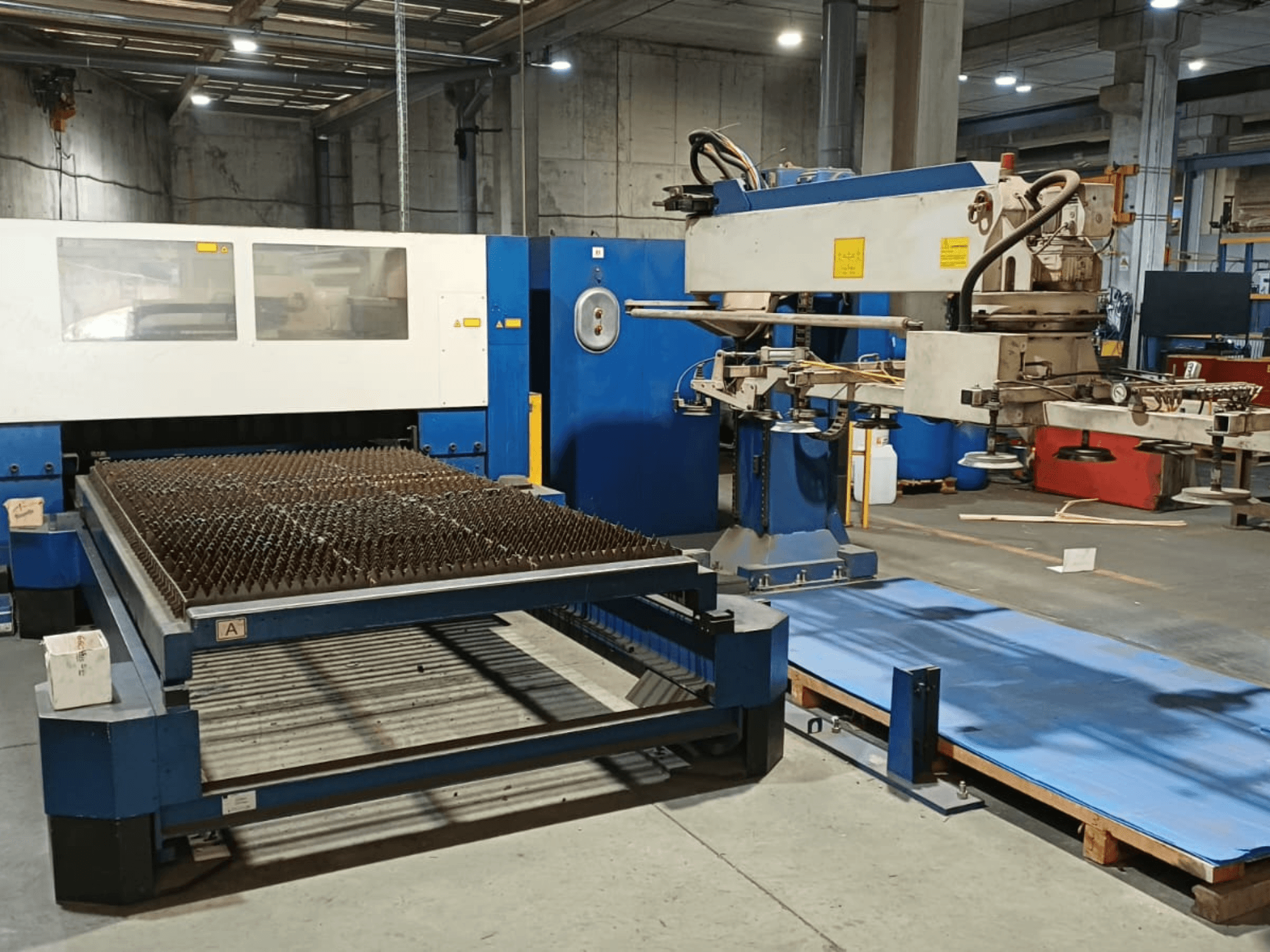 Laserový řezací stroj TRUMPF TRUMATIC L 3030 s CO₂ laserem, boční pohled, na kterém je vidět řezací stůl s hrbolatým povrchem a robotické rameno.