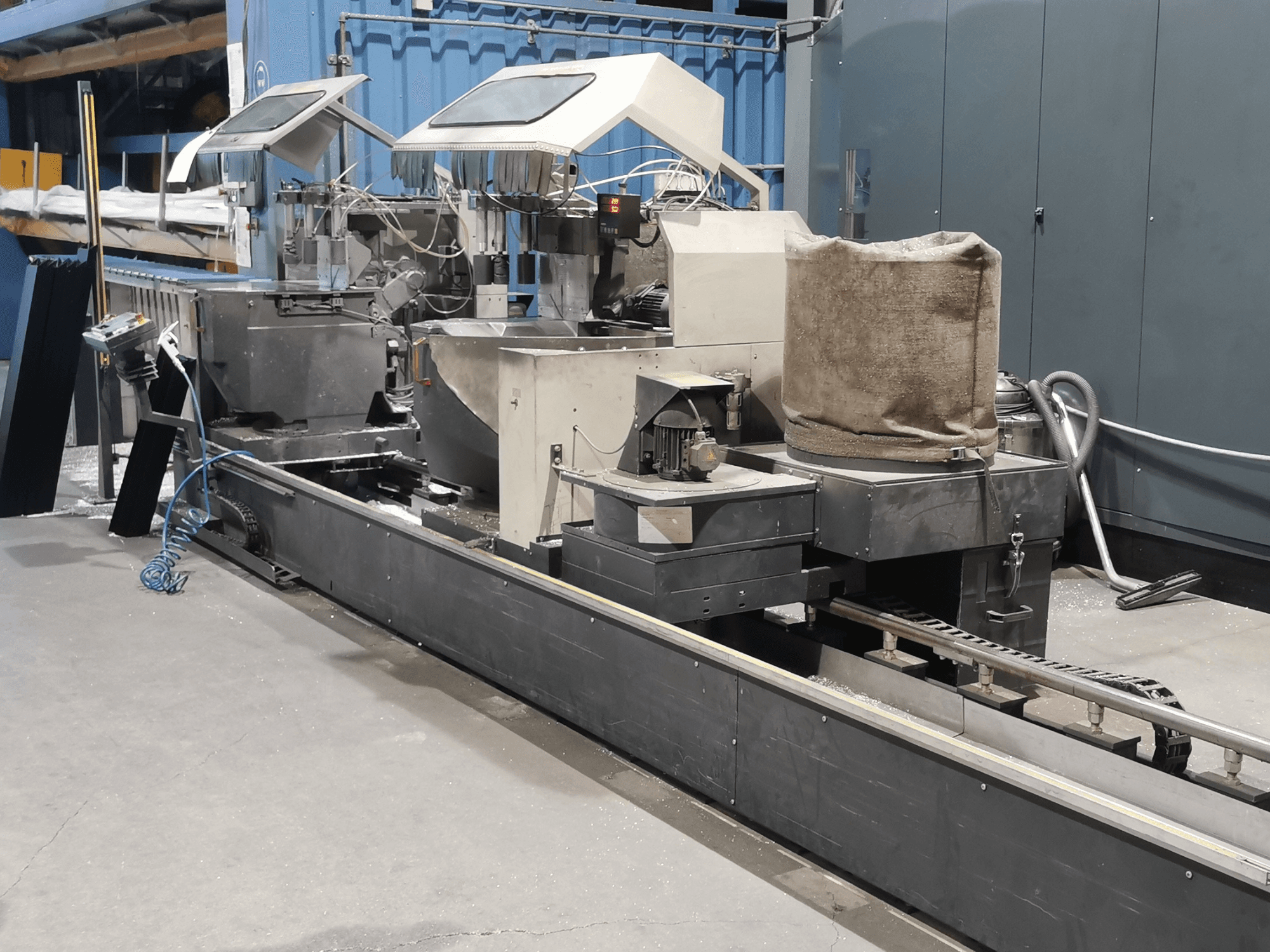 Průmyslový zpracovatelský stroj z bočního pohledu, se zásobníky, dopravníkovým pásem a ovládacím panelem v továrním prostředí.