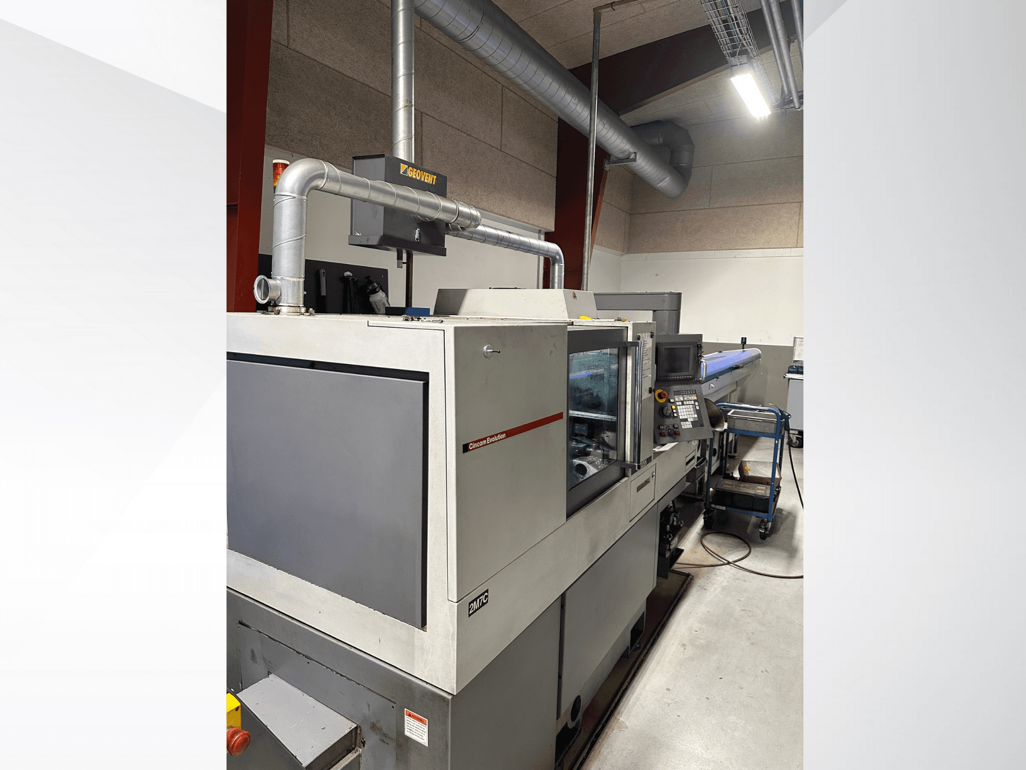 Průmyslové stroje značky MVC, boční pohled s viditelným ovládacím panelem a potrubím, zasazené do továrního prostředí.