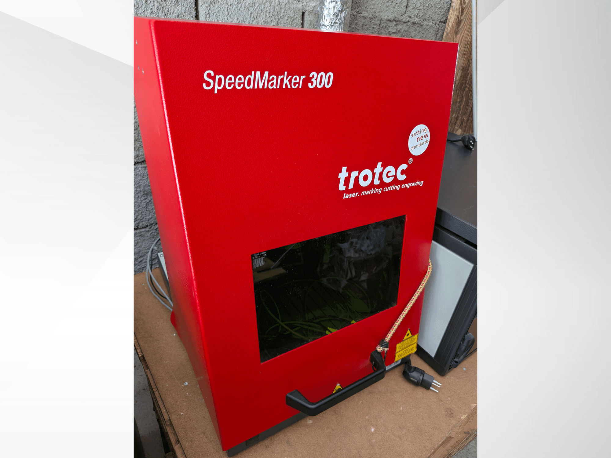 Červený laserový značkovací stroj Trotec SpeedMarker 300 v čelním pohledu, s velkým průzorem a přípojkou napájení.