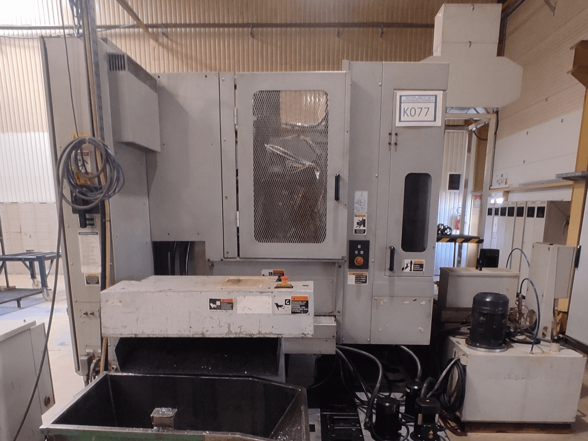 Šedý průmyslový CNC stroj s velkým pracovním stolem, boční pohled, s ovládacím panelem a zásobníkem chladicí kapaliny, model K077.